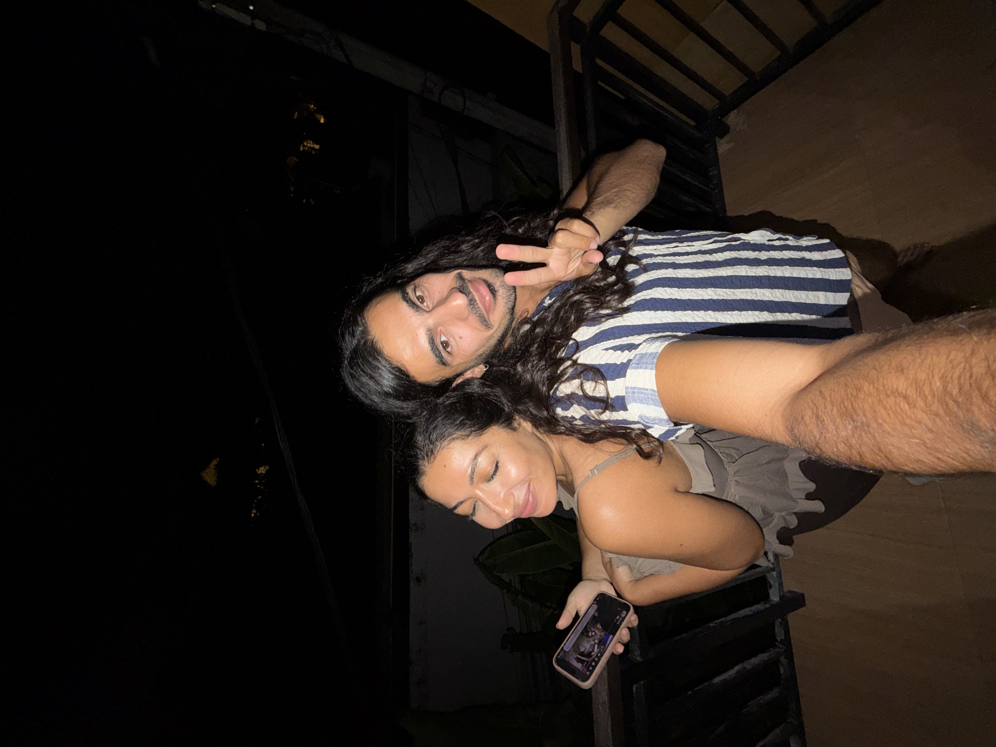 This is a nighttime selfie featuring two individuals, a man and a woman, posing on what appears to be an outdoor balcony or terrace in North Kuta, Indonesia. The image is tilted, indicating a dynamic capture, and the background is predominantly dark, confirming the late hour.
The man, positioned towards the top of the frame, has long, dark, curly hair and a beard. He is smiling broadly at the camera and making a peace sign with his right hand. He wears a blue and white striped short-sleeved shirt. His left arm, showing visible body hair, extends into the dark background.
Below and slightly in front of him is the woman, also with dark, curly hair. She is looking down and to her right with a gentle, relaxed smile, her eyes appearing partially closed. She is wearing a light-colored, possibly beige or grey, sleeveless top with thin straps. In her left hand, she holds a dark-colored smartphone, its screen illuminated and displaying an image, though specific details on the screen are indistinct. Her right arm is bent, resting near a dark metal railing that runs along the edge of the balcony.
The scene is illuminated by artificial light, casting soft glows on the subjects. The immediate surroundings include the dark metal railing and a light brown, possibly tiled, floor visible to the right. The background is largely dark and indistinct, with faint suggestions of foliage or distant structures, consistent with an outdoor nighttime setting. The overall mood conveyed is relaxed and amicable. No discernible text is visible within the image.