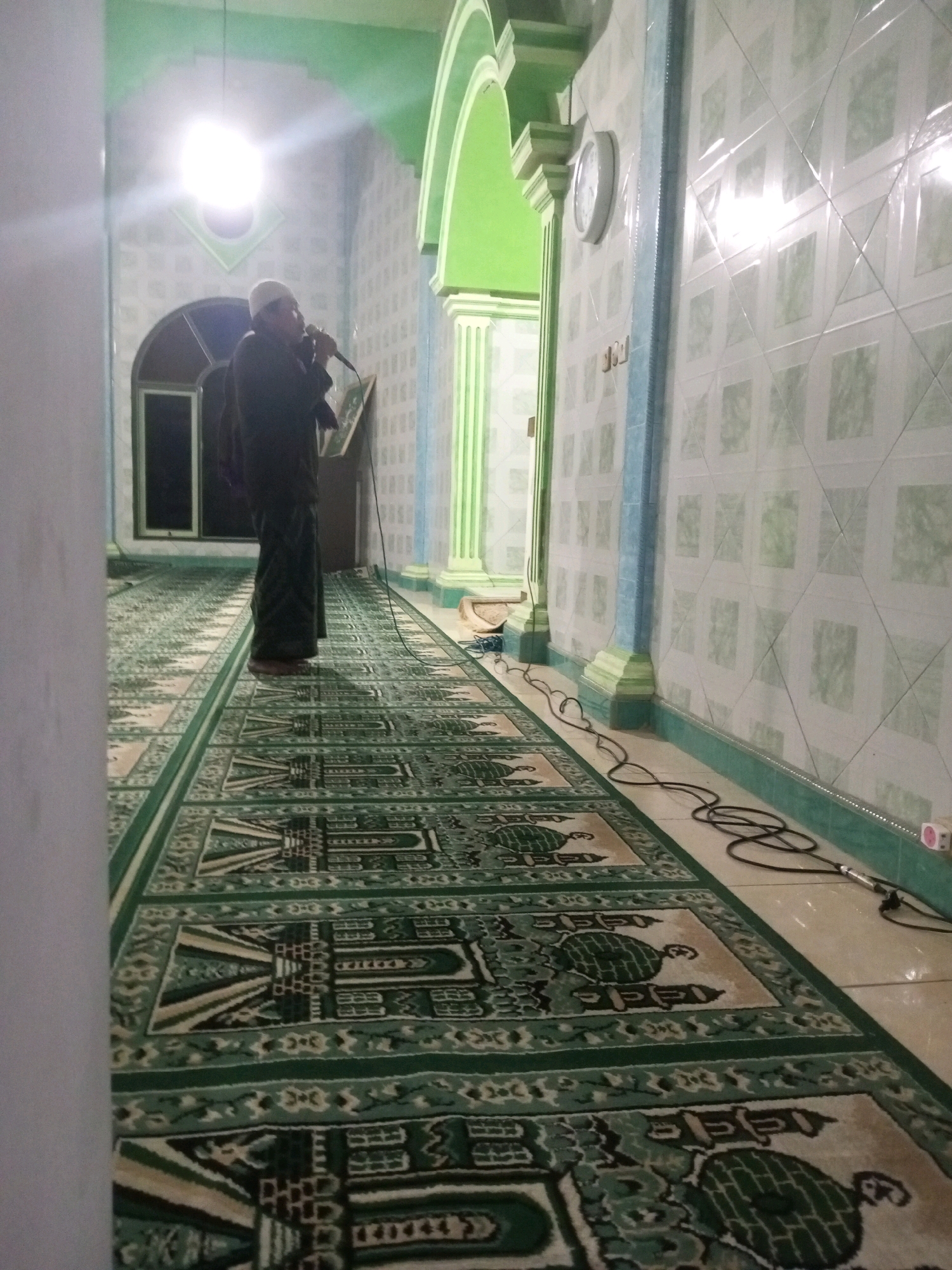 In the city of Andongsili, Indonesia, a man stands on a patterned carpet inside a room, holding a microphone and speaking. He is wearing a white skullcap and dark clothing. The room features walls covered in square-patterned tiles and green architectural elements. A clock hangs on one wall, and a large, bright light fixture is visible overhead. Cables snake across the floor, connected to what appears to be audio equipment. The carpet features a repeating pattern of mosque-like structures and floral motifs in green and cream.