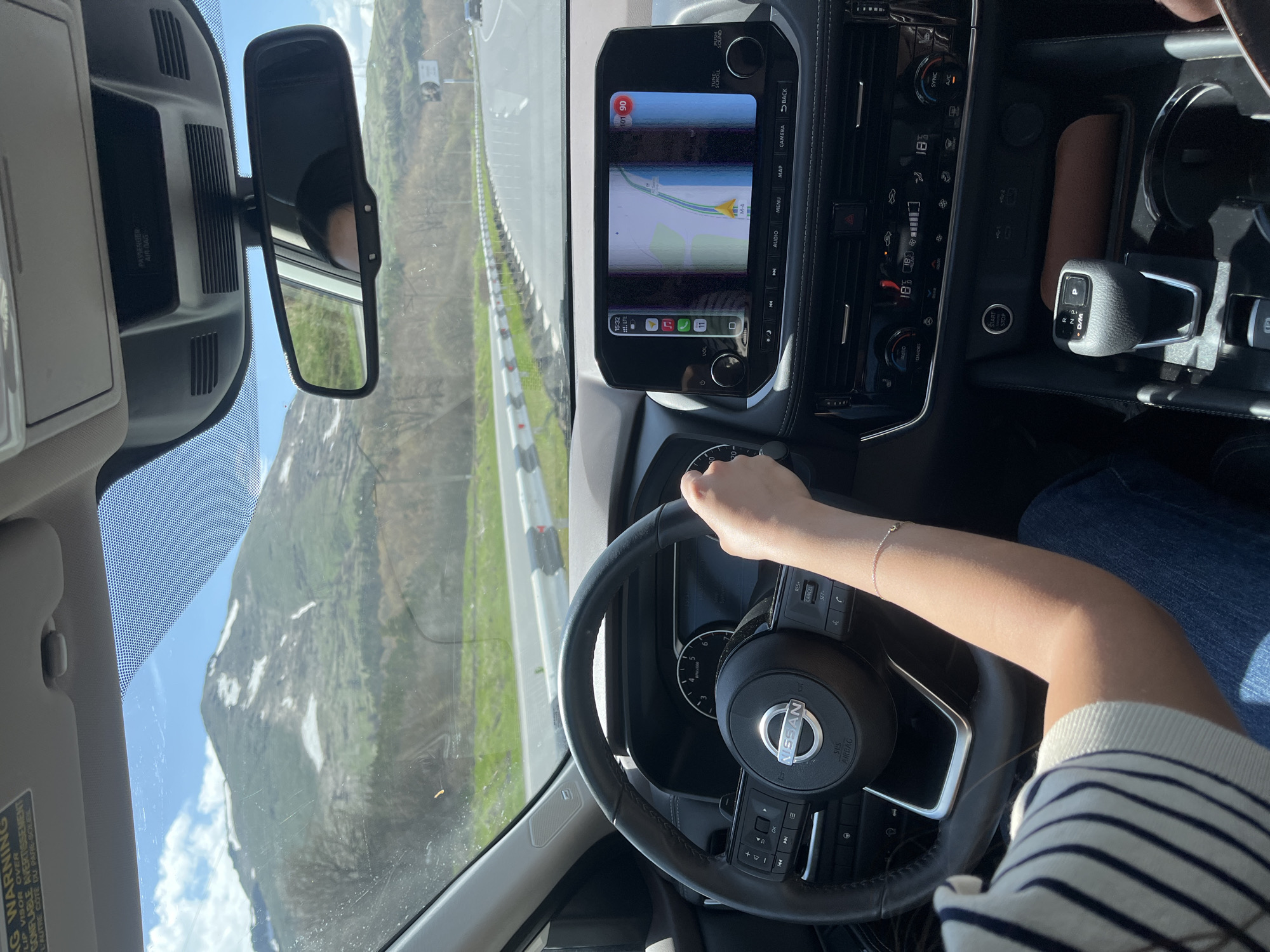 The image captures an interior view from the driver's perspective of a Nissan vehicle traveling on a road in Tsovagyugh, Armenia. The dashboard shows a map displaying the M-4 route. The driver's hand is on the steering wheel, and the gear shift is in park. The exterior shows a mountainous landscape with clear skies.