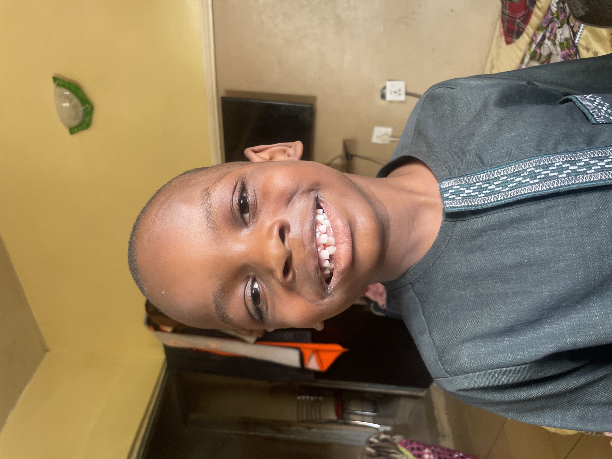 A boy with a shaved head is smiling at the camera. He is wearing a dark grey shirt with decorative trim around the neck and shoulder. The background shows a yellow wall with a green wall light fixture and an electrical outlet. A television is visible behind the boy. There is no information that places the image in Sabon Tasha, Nigeria.