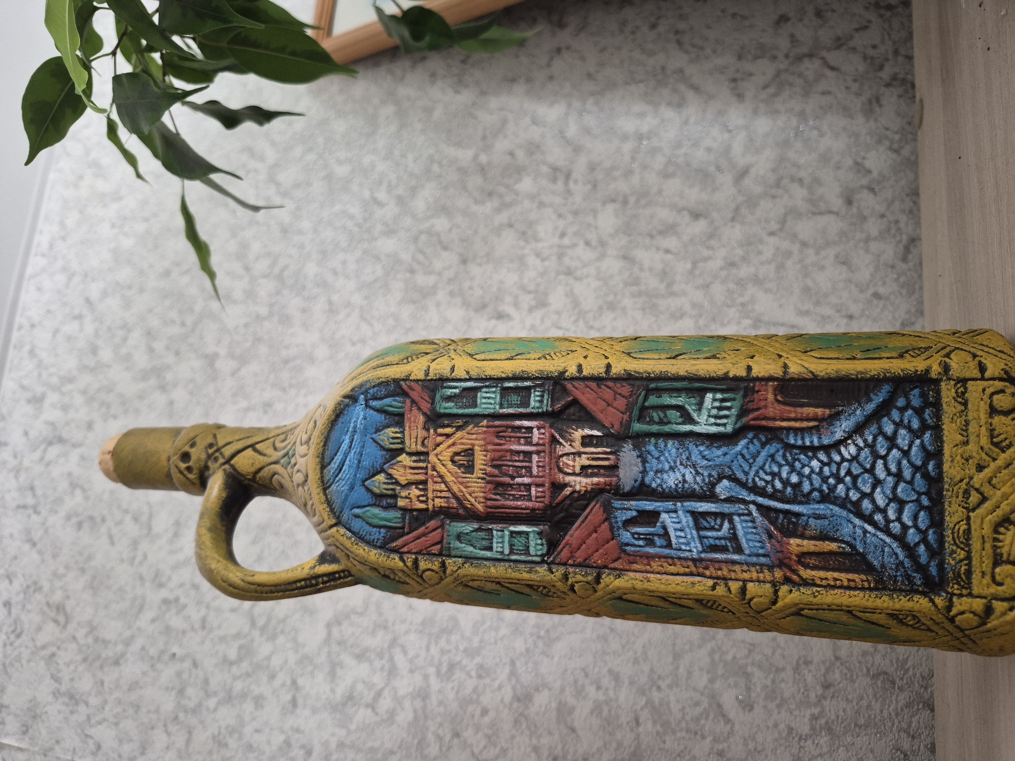The image shows a decorative ceramic bottle lying horizontally on a surface. The bottle is predominantly yellow with carved and painted details in blue, red, and green. The design includes architectural elements and what appears to be a depiction of a dragon. A handle is attached to the neck of the bottle. A small plant is visible in the upper left corner. The background is a mottled gray and white surface, possibly a countertop or wall.