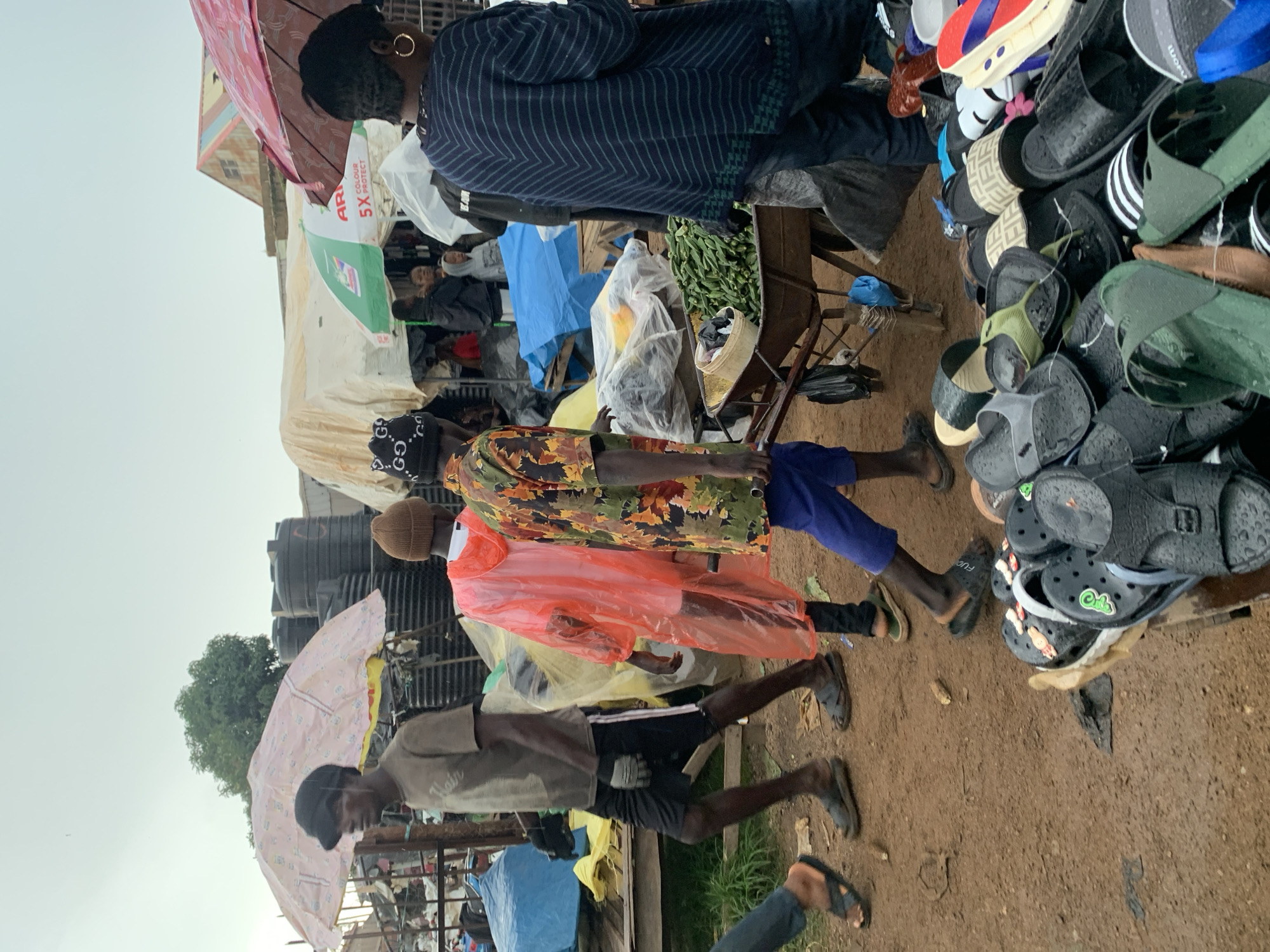 The image depicts a bustling outdoor market scene in Gura Top, Nigeria, under an overcast sky. The ground is dirt, and there are various stalls and makeshift structures covered with tarpaulins and umbrellas. Several people are visible, engaged in shopping or selling activities.
In the foreground, a collection of footwear, primarily sandals and flip-flops, is piled up on a wooden surface, suggesting a shoe stall. A wheelbarrow filled with green vegetables, possibly okra, is also prominent, with a white bucket containing various items inside.
Several individuals are walking through the market. One person in the mid-ground wears a bright orange plastic raincoat and black leggings. To their left, a man in a brown t-shirt and black shorts walks with a slight stoop, possibly carrying something unseen. Another man, to the right of the person in the orange raincoat, wears a colorful patterned shirt and blue shorts. Further back, other market-goers are partially visible, including a woman in a striped sweater and a person in a black beanie with white lettering.
The overall impression is one of a lively, functional marketplace with a diverse array of goods and people going about their daily business. The presence of umbrellas and the overcast sky suggest potential for rain or a humid environment.