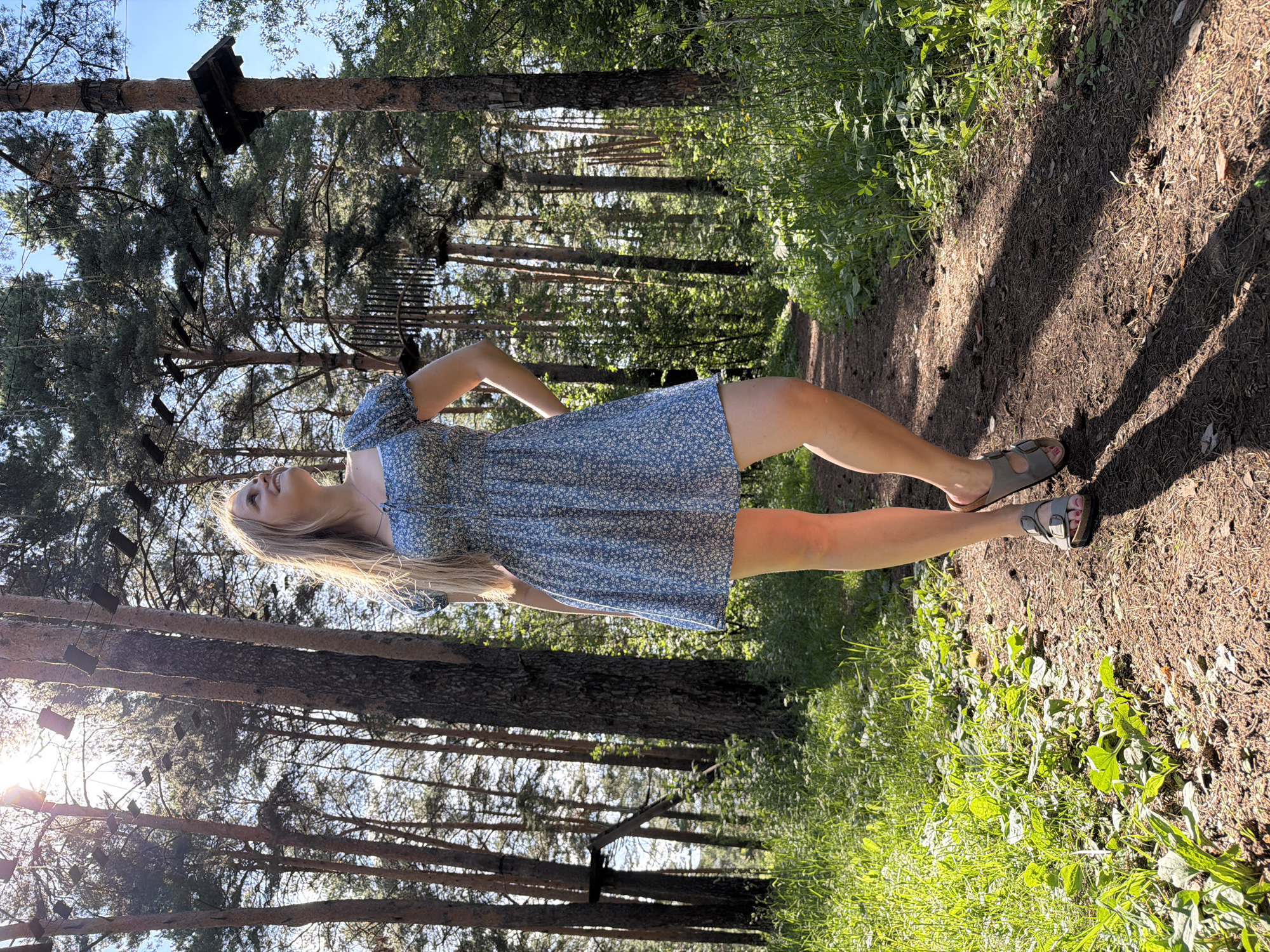 In Izhevsk, Russia, on a sunny day, a young woman with blonde hair stands on a dirt path in a forest. She is wearing a short, blue floral dress and gray sandals. The path is lined with lush green grass and tall pine trees. In the background, rope courses are visible, suspended between the trees, indicating this is likely an adventure park or recreational area. The sun casts long shadows across the path.