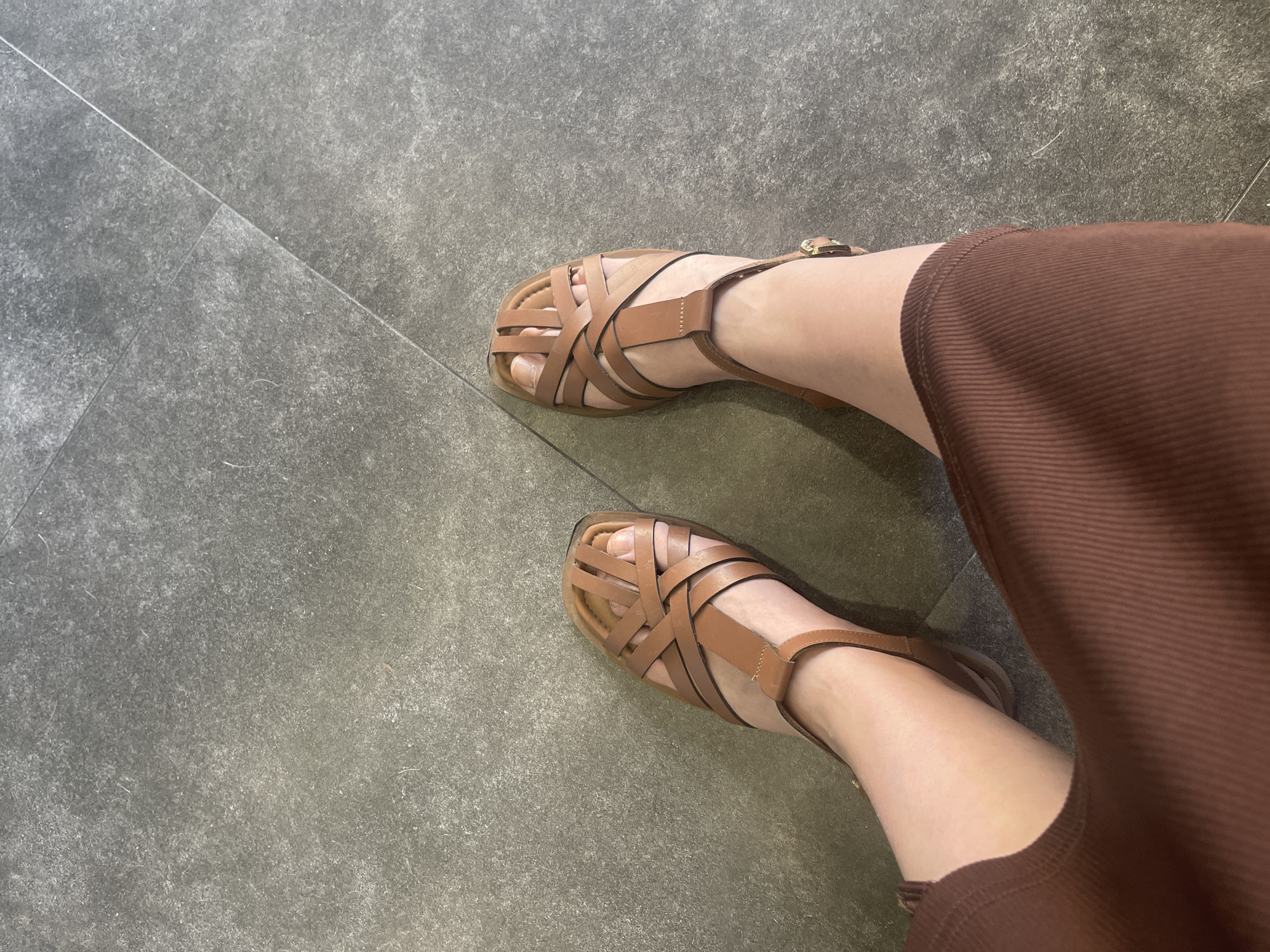 This media file is a close-up, high-angle shot of a person's legs and feet, wearing brown woven sandals. The person is also wearing a brown, ribbed skirt or dress. The footwear is a flat sandal with multiple straps that crisscross over the toes and an ankle strap with a buckle closure. The scene is set on a gray tiled floor, with visible grout lines creating a geometric pattern. The lighting suggests it is daytime, possibly indoors given the consistent illumination and lack of shadows. No specific location cues or environmental details, such as weather or time of day beyond daytime, are apparent. The image focuses solely on the fashion items and the lower portion of the person's body. There is no visible text within the image. The image captures a simple, everyday moment, likely a personal photograph documenting an outfit.