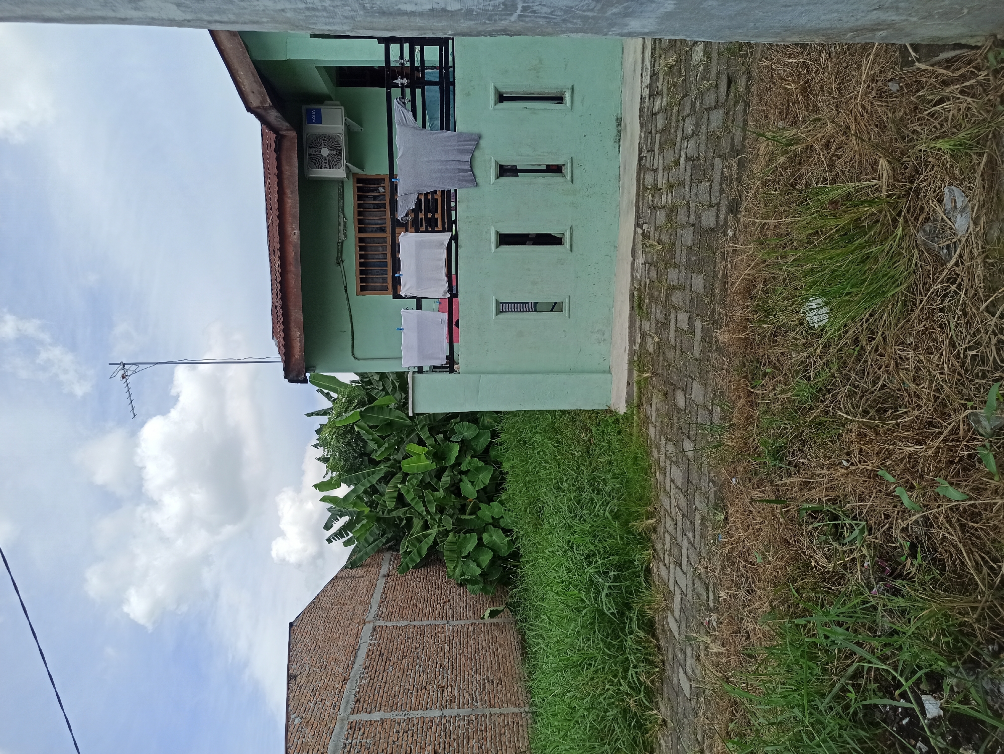 In the City of Medan, Indonesia, this image captures a scene with a light green, two-story residential building situated to the right. The building features a rusty corrugated metal roof and an air conditioning unit mounted on the exterior wall. Laundry, including a grey t-shirt and two white shirts, is hung on a railing on the upper floor. Below the railing, there are four narrow, rectangular window openings. To the left of the building, a brick wall is visible, partially covered by lush green banana plants. The foreground is a mix of dry, brown grass and some green grass, with a paved walkway made of interlocking stone bricks running across the middle ground. The sky above is cloudy with patches of blue.