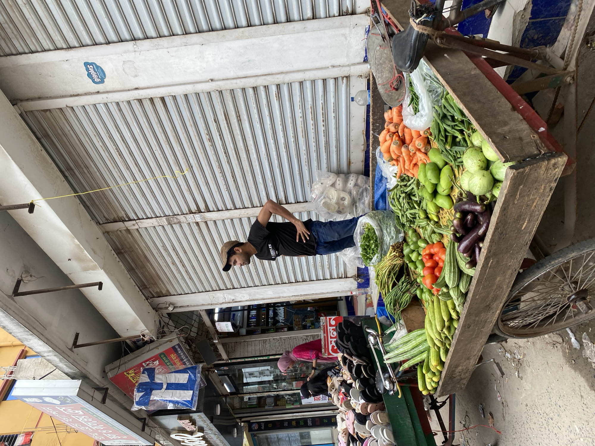 A vendor stands next to a wooden vegetable cart on a sidewalk in Dhaka, Bangladesh. The cart is loaded with carrots, long beans, green vegetables, tomatoes, eggplants, and gourds. The vendor is a dark-haired man wearing a black t-shirt, blue jeans, and a baseball cap. The sidewalk is lined with storefronts, including a shoe store and other shops. Above, a corrugated metal awning provides shelter. A Finnish flag hangs from one of the storefronts. The scene captures a typical marketplace in Dhaka.