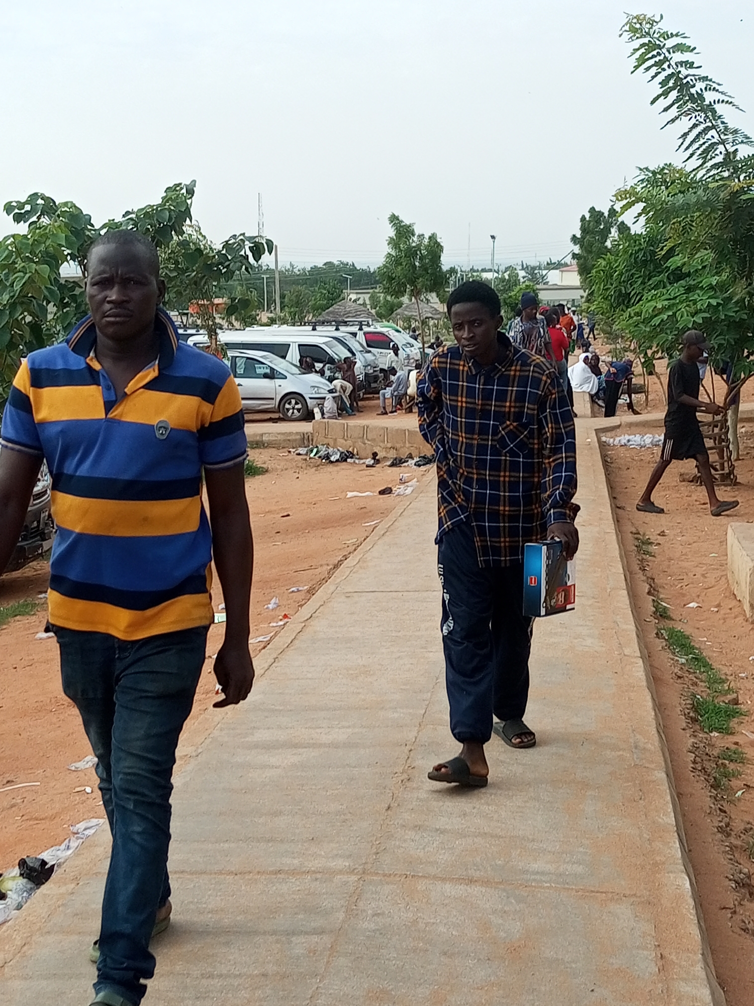 In Gombe, Nigeria, a man in a blue and yellow striped polo shirt and blue jeans walks on a paved path. He is looking directly at the camera with a neutral expression. To his right, another man wearing a blue and yellow plaid shirt and dark pants walks towards the viewer, carrying a box. In the background, several white vans are parked in a lot. The ground is sandy and there are trees and other foliage scattered throughout the scene. The sky is overcast.