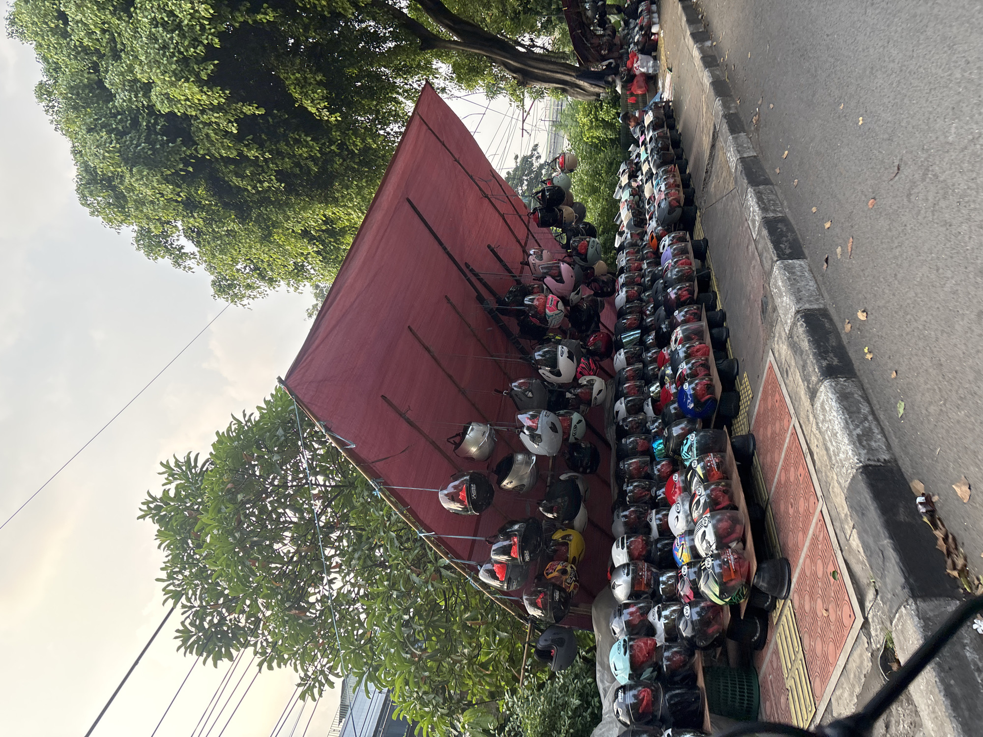This media file captures an outdoor stall selling a large collection of motorcycle helmets. The scene is set on a sidewalk next to a paved road, with lush green trees and foliage providing a backdrop. A large, reddish-brown tarp is stretched over a simple frame, creating a shaded area for the display.
The primary objects are hundreds of motorcycle helmets, neatly arranged on racks and shelves. Some helmets are hung from the tarp's frame, while others are stacked in rows on tables and on the ground. They come in a variety of colors, designs, and types, including full-face helmets, open-face helmets, and what appear to be children's helmets. The sheer volume suggests a retail or rental operation.
The setting appears to be a street in South Jakarta, Indonesia, indicated by the presence of utility lines and the overall urban environment. The time of day seems to be late afternoon or early evening, as suggested by the quality of light, which casts long shadows and has a warm hue. The sky is partly cloudy. No people are clearly visible in the foreground, though some figures might be obscured by the helmets or out of frame. There is no visible text in the image. The overall impression is a bustling marketplace or a roadside vendor catering to motorcycle riders.