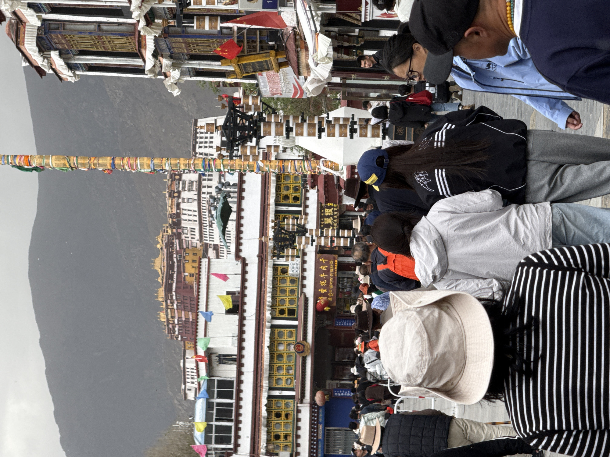 The image shows a crowd of people in front of a large building complex. The complex has white walls, multiple floors, and windows with yellow frames. Prayer flags hang from a tall pole in front of the buildings. The background features a mountain under an overcast sky. This image appears to be taken in the Chengguan District in China, as there are traditional buildings there with white walls and windows with yellow frames. The people in the foreground appear to be tourists observing the architecture.