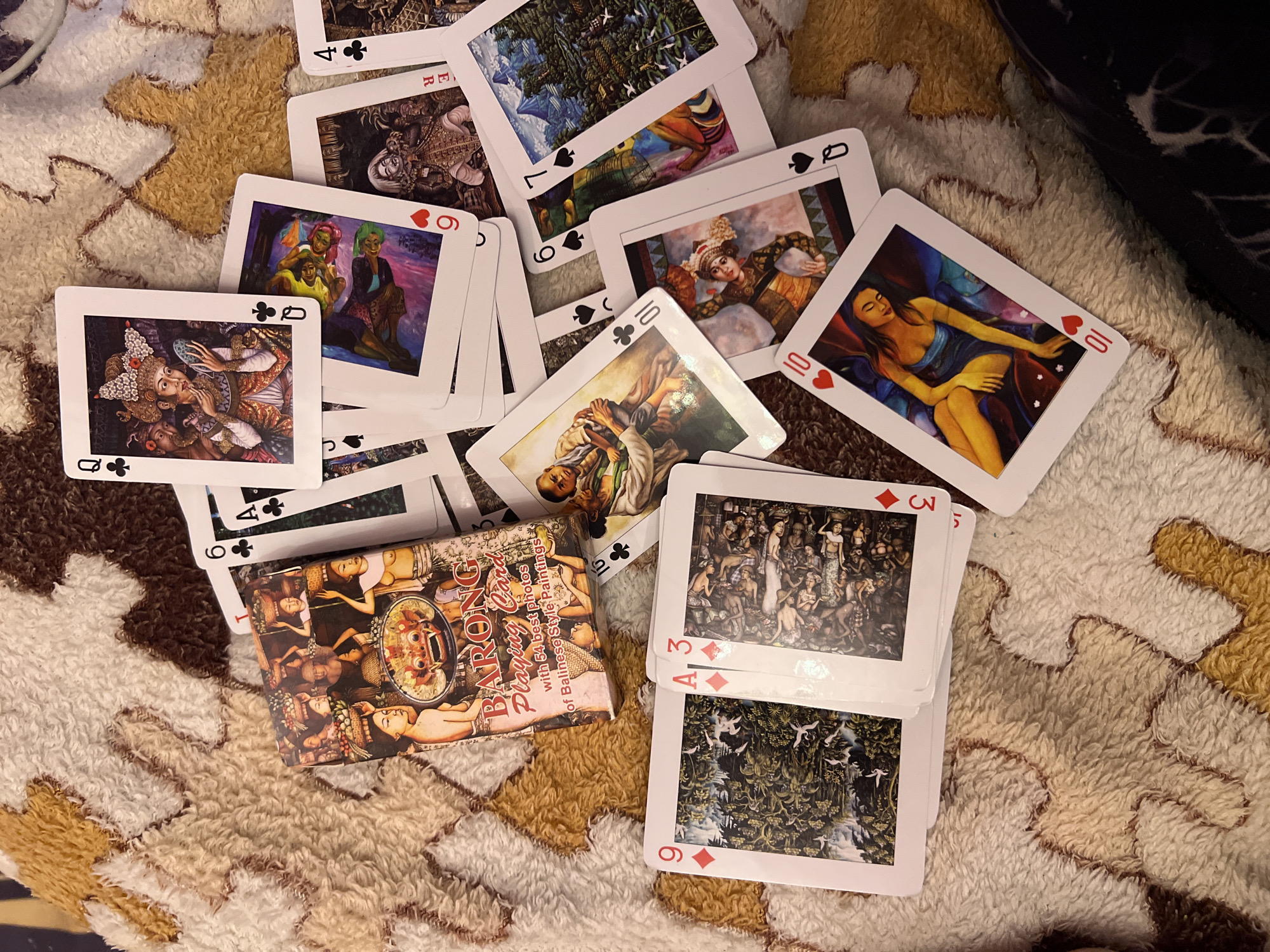 The image shows a close-up, top-down view of a pile of playing cards spread across a textured, fluffy blanket with a pattern of brown, tan, and cream colors. Some of the cards are face up, revealing intricate, colorful artwork depicting figures and scenes, many in a style that appears to be Balinese. One card, a Queen of Clubs, features a detailed portrait of a woman in ornate headdress and traditional clothing applying makeup. Another card, a 10 of Hearts, displays a woman in a blue dress. The deck box, labeled "BARONG Playing Card with 54 best photos of Balinese Style Paintings," is partially visible, positioned in the upper left quadrant of the image. Several other cards are visible, including a 9 of Diamonds showing a landscape with birds, a 3 of Diamonds with a dense, crowded scene of figures, a 6 of Clubs, a 7 of Spades, a 10 of Spades, and a King of Spades. The cards are fanned out and overlapping, suggesting they have been used or are being displayed. The blanket appears to be a throw or rug, contributing to a casual indoor setting. There is no indication of the city of Krasnoe-na-Volge, Russia, in the image itself or its contents.
