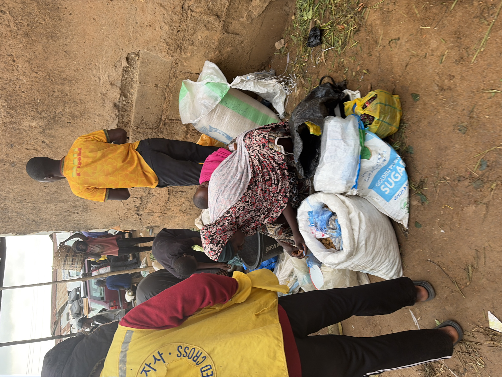 In Gura Top, Nigeria, a group of individuals are engaged in what appears to be a distribution or collection of goods. In the foreground, a person wearing a bright yellow vest with "RED CROSS" printed on it in Korean is partially visible. Adjacent to them, a person in a red and black patterned headscarf and dress is seated, sorting through items in a white sack. Other individuals are bent over or standing around piles of sacks, some of which contain food items. A man in a yellow t-shirt stands nearby, observing the scene. The environment is dusty and appears to be an outdoor market or communal area, with a rough-textured wall as a backdrop. Sacks of what seem to be grains and sugar are prominent, along with other packaged goods. The overall impression is one of a community effort involving aid distribution or local commerce.