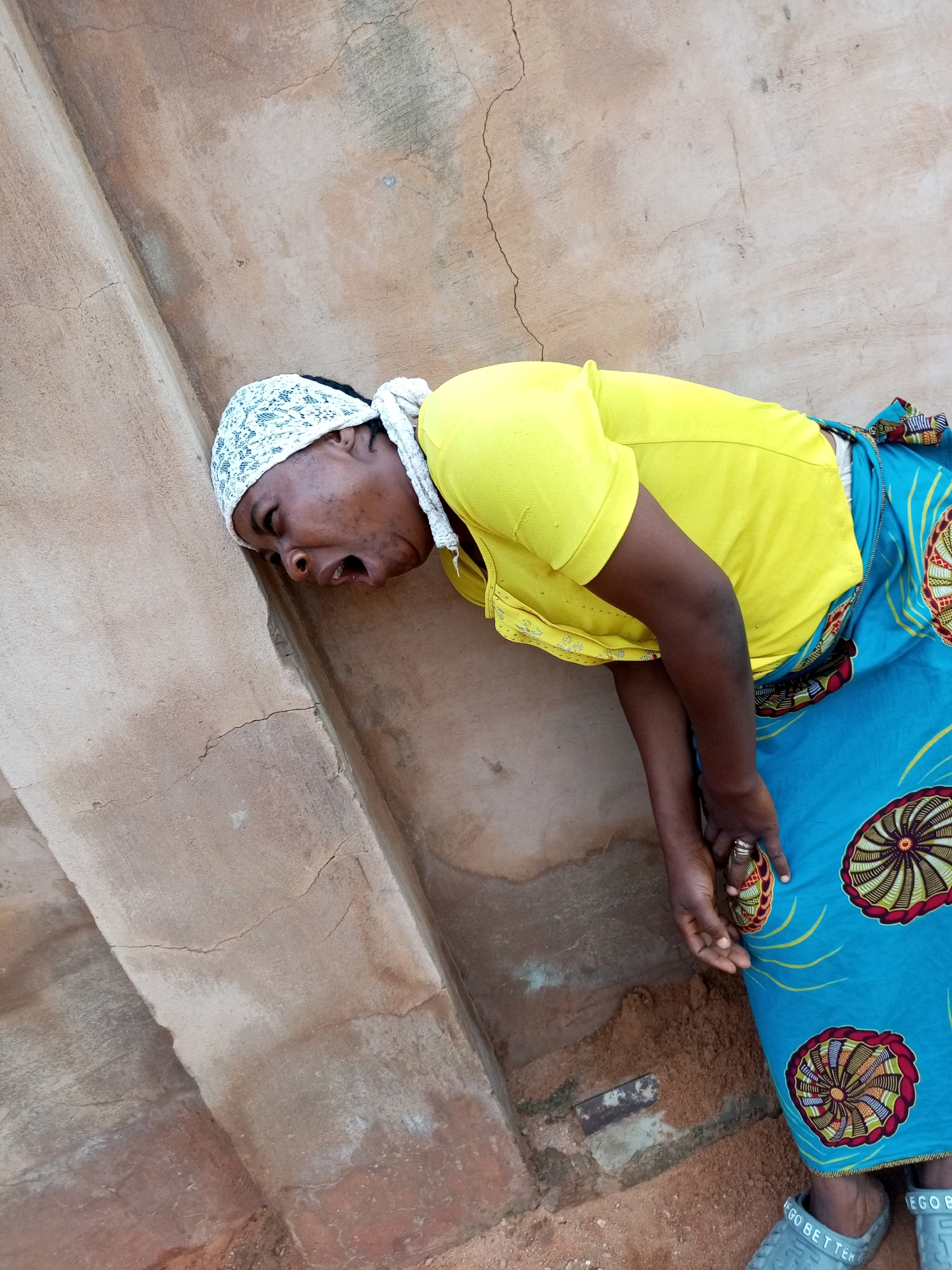 A person wearing a yellow shirt and a blue and patterned skirt is leaning against a cracked, tan-colored concrete wall. They have a white headscarf and their mouth is open in a cry or shout. Their right hand is holding a small, colorful object near their chest. The person is barefoot and wearing grey slippers. The scene appears to be outdoors, with dirt and debris on the ground. The wall has visible cracks and stains, indicating it is weathered. The lighting suggests it is daytime. The location is the city of Tempure, Nigeria.