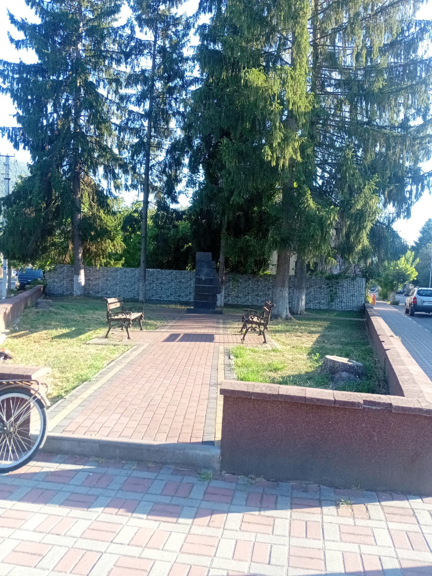 The image shows a public park or square in Tiachiv, Ukraine. In the foreground, there is a paved pathway made of red and gray interlocking bricks, with a curb separating it from a grassy area. To the left, a portion of a bicycle wheel is visible. Further into the park, two wrought-iron benches are placed on the grass. Behind the benches, a low stone wall runs horizontally, and beyond that, a monument or obelisk is situated. Large evergreen trees dominate the background, casting shadows on the ground. A few parked cars are visible in the distance on a street adjacent to the park. The scene appears to be a quiet, well-maintained outdoor space bathed in sunlight.