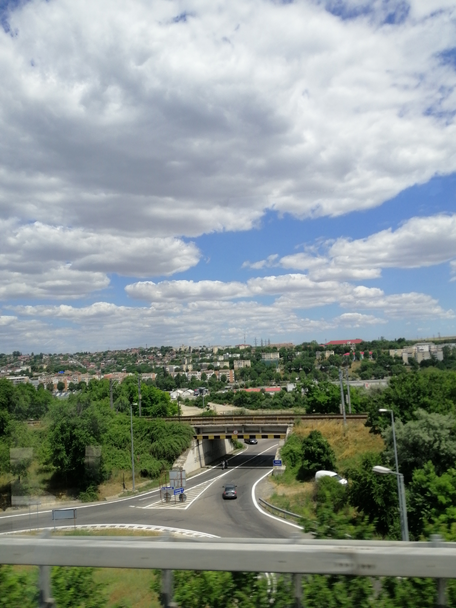 The media file depicts a highway scene on a cloudy day with a backdrop of a distant cityscape. The foreground features a gray metal railing, blurred by motion, hinting at a moving vehicle. Below the railing, lush green foliage is visible. The main focus is a multi-lane highway curving through a landscape.
Several cars are seen on the road, with one black car driving through an underpass and another dark gray car visible on the curved section of the highway. An overhead bridge spans the highway, and a light pole with a lamp is situated on the right side of the road. To the left of the road, there are some informational signs, one with a blue and white symbol and another blank sign.
In the middle ground, dense green trees line the highway and the slopes. The background reveals a sprawling city with numerous buildings of varying sizes and colors, nestled on rolling hills. The sky is filled with large, fluffy white clouds interspersed with patches of blue, suggesting it is daytime. The overall impression is a typical suburban or semi-urban highway environment under a partly cloudy sky. There is no discernible text in the image that can be read clearly.