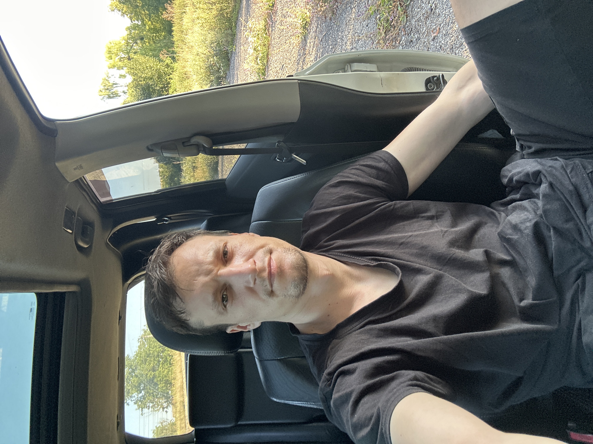 A man is sitting in the driver's seat of a car with the door open, looking at the camera. The car appears to be parked on a gravel road in a rural area. The background shows a field of tall grass and trees. The man is wearing a black t-shirt and shorts. The sun is setting, casting a warm glow on the scene. The location is Makiv, Ukraine.