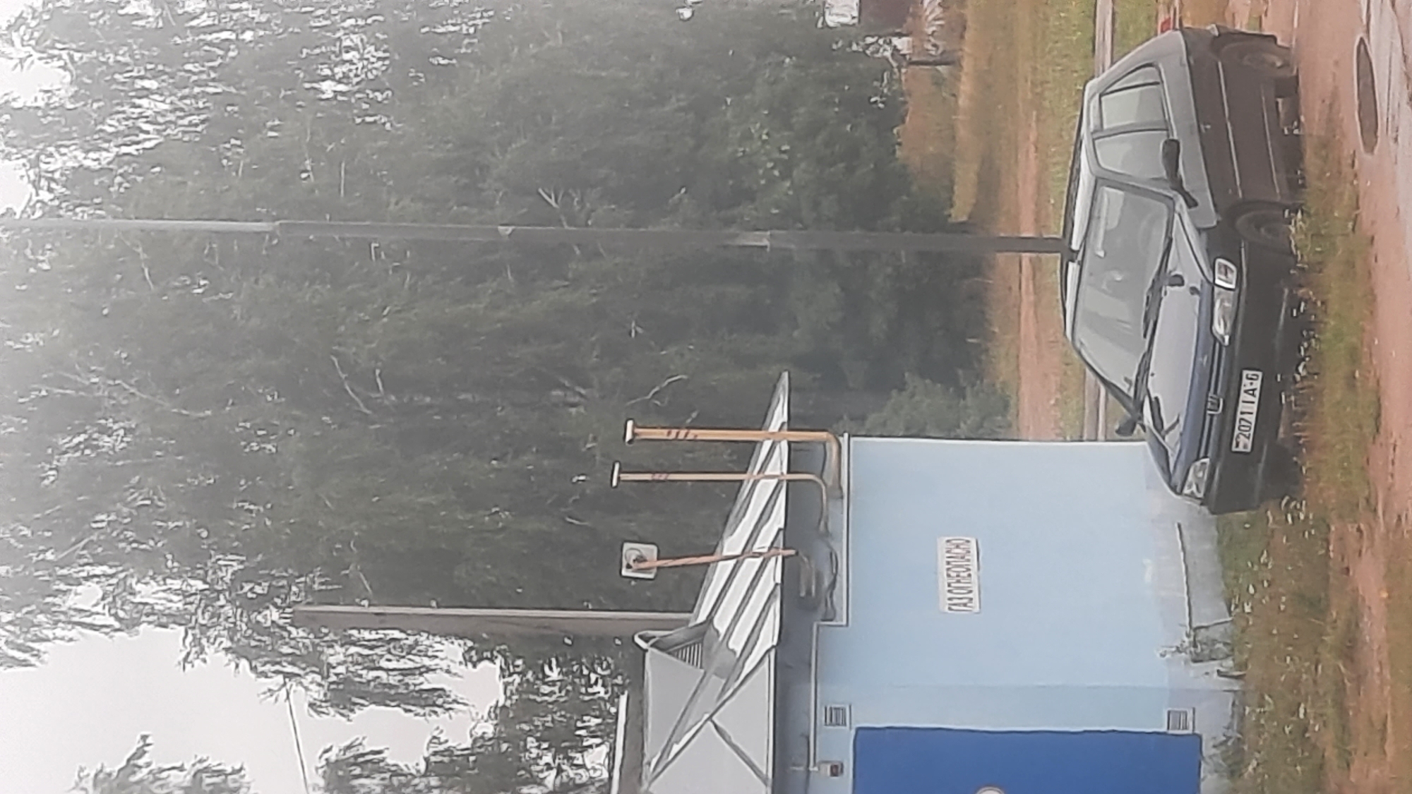 A dark blue, four-door hatchback is parked on a gravel and grass area in front of a light blue, rectangular building with a grey, pitched roof. The building has a blue door on the left side and a sign on the front that reads "ГАЗ ОТНЕОПАСНО". Two yellow exhaust pipes emerge from the roof of the building, bent at a 90-degree angle. A single, thin, dark pole stands vertically behind the building and car, extending upwards. Dense green foliage, appearing to be trees, forms the background. The sky is overcast and a light haze is visible. The license plate of the car reads "20711A-6".