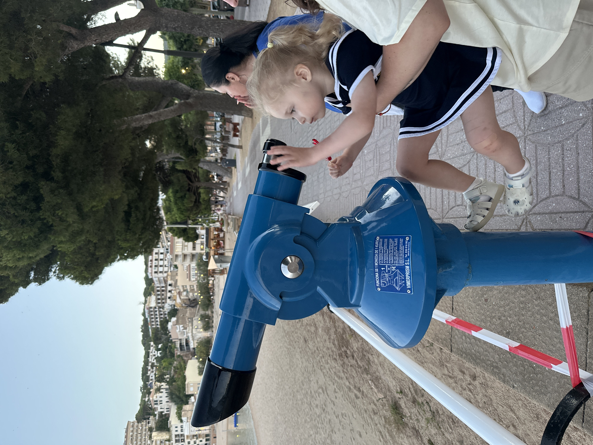 A young girl with blonde pigtails, wearing a black and white striped dress and white sandals, is looking through a large blue coin-operated telescope on a beach promenade. She is being held by an adult whose arm is visible. In the background, a woman with dark hair is visible looking towards the sea. The promenade features a paved walkway and a sandy beach with the sea in the distance. Buildings line the hillside behind the beach, with trees and streetlights visible along the promenade. The sky is clear and a light blue, indicating it is either early morning or late afternoon. Signage on the telescope indicates it requires one euro to operate and provides instructions in multiple languages. The overall scene suggests a recreational outing at a coastal tourist destination. The city of Sant Feliu de Guíxols, Spain is implied by the signage and the architectural style of the buildings, consistent with coastal towns in this region.