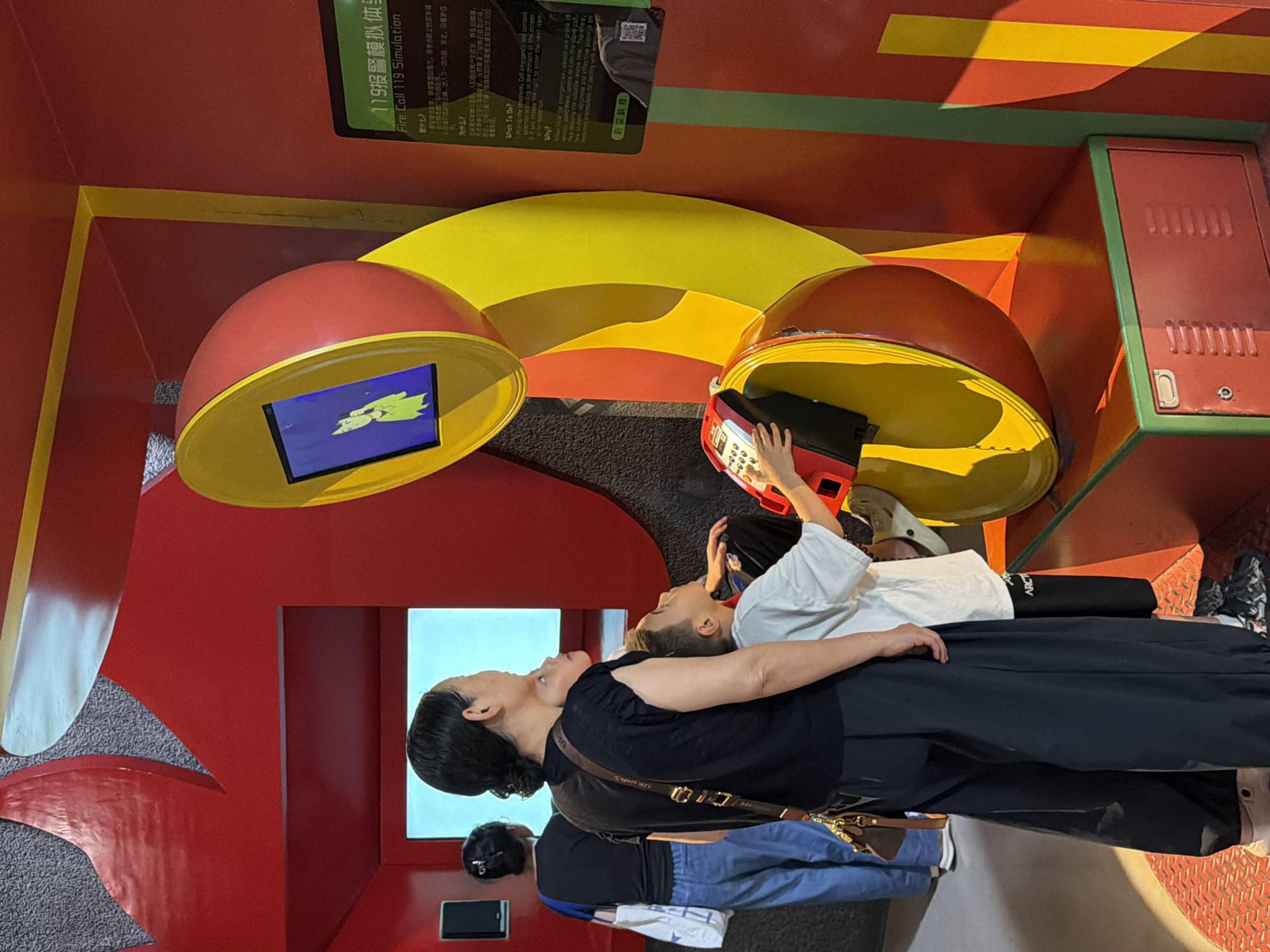 In the city of Jiangbei City Street, China, a simulated fire call exhibit is on display. The exhibit features a large, stylized red and yellow telephone receiver that is cut in half and appears to be an interactive screen. A woman, her back to the camera, stands with two children. One child, a boy, is actively engaged with a red and gray phone handset at the base of the exhibit, his hand on the keypad. The other child, also a boy, stands between the woman and the interactive phone, looking up at the screen. In the background, another woman wearing a blue shirt and carrying a white bag walks away from the exhibit. The environment is brightly colored, with red walls and yellow and green accents, suggesting an educational or recreational space, possibly a museum or exhibition hall. A sign with Chinese characters and "Fire Call 119 Simulation" in English is visible on the right side of the image.
