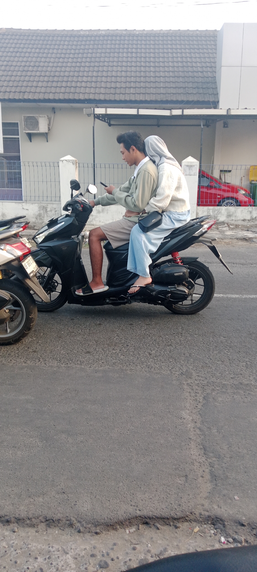 Two individuals, a male driver and a female passenger, are riding a black scooter on an asphalt road. The male is wearing a light-colored long-sleeved shirt and brown shorts, and is holding a smartphone. The female is wearing a white sweater and light blue pants, with a light-colored hijab covering her hair. She has her left arm around the male's waist. A black pouch is slung across her body. They are positioned in the foreground, with a fence and buildings in the background. A red car is visible behind the fence. The scene appears to be in Maguwoharjo, Indonesia, likely on a street with residential buildings.