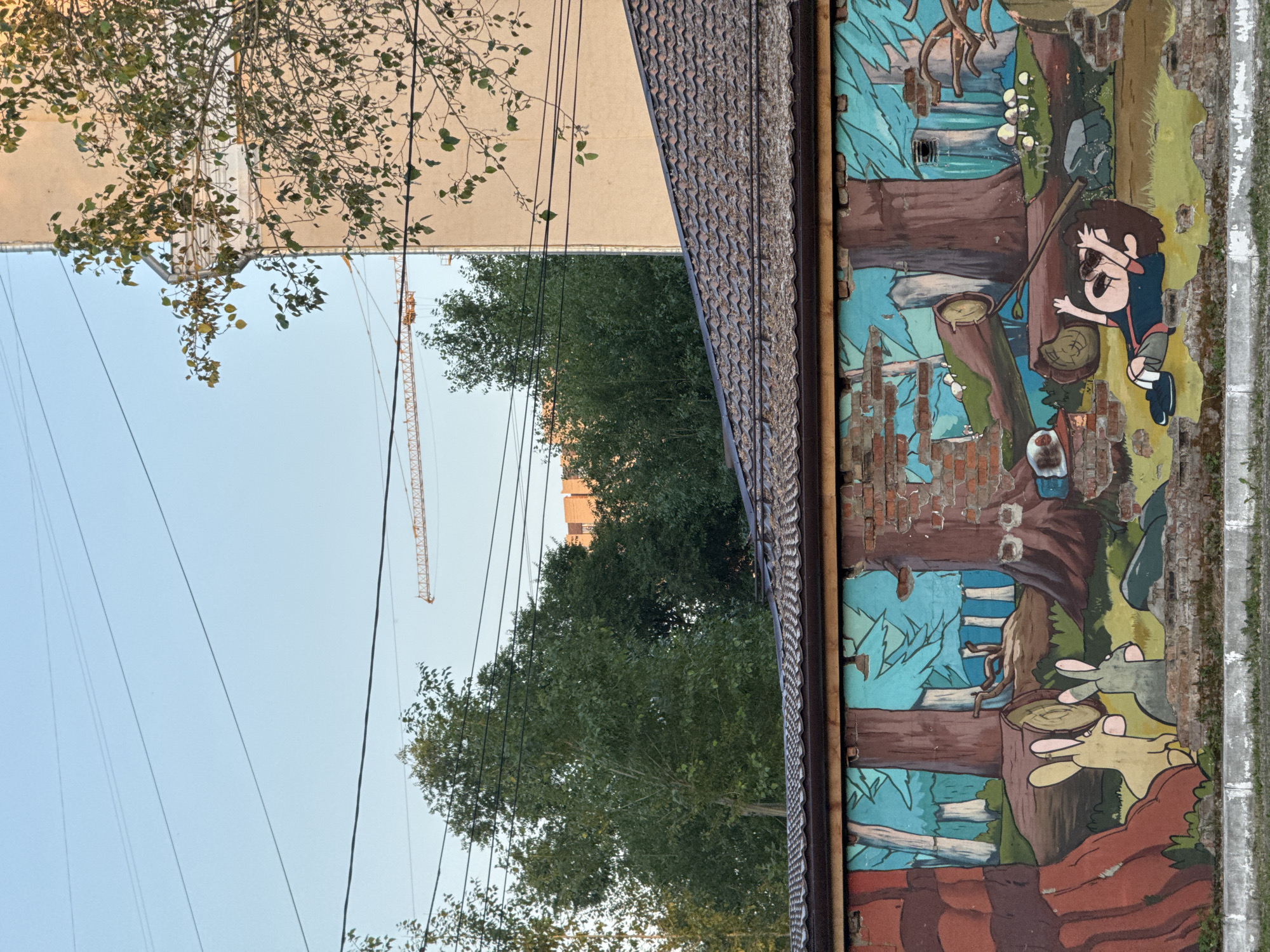 The image shows a mural painted on the side of a building in Krasnoyarsk, Russia. The mural depicts a cartoon scene of woodland creatures, including two rabbits, and a young boy. The boy appears to be looking upwards with a surprised or fearful expression. He is wearing a blue shirt and grey shorts. The rabbits are depicted in profile, with large ears and pink noses. The background of the mural features trees, logs, and mushrooms, rendered in a somewhat abstract style with a palette of blues, greens, and browns. The mural is painted on a brick wall that shows signs of damage and wear, with some bricks missing.
To the left of the mural, a building with a beige facade is visible, with a tree in the foreground casting shadows. Above the tree and building, a pale blue sky is visible, with several power lines running across it. A tall construction crane with a yellow lattice structure is also visible in the distance, suggesting ongoing construction in the area. The lighting suggests it is daytime, possibly late afternoon, with soft light. The overall scene combines urban elements with a whimsical mural, creating a somewhat surreal juxtaposition. No text is visible in the image.