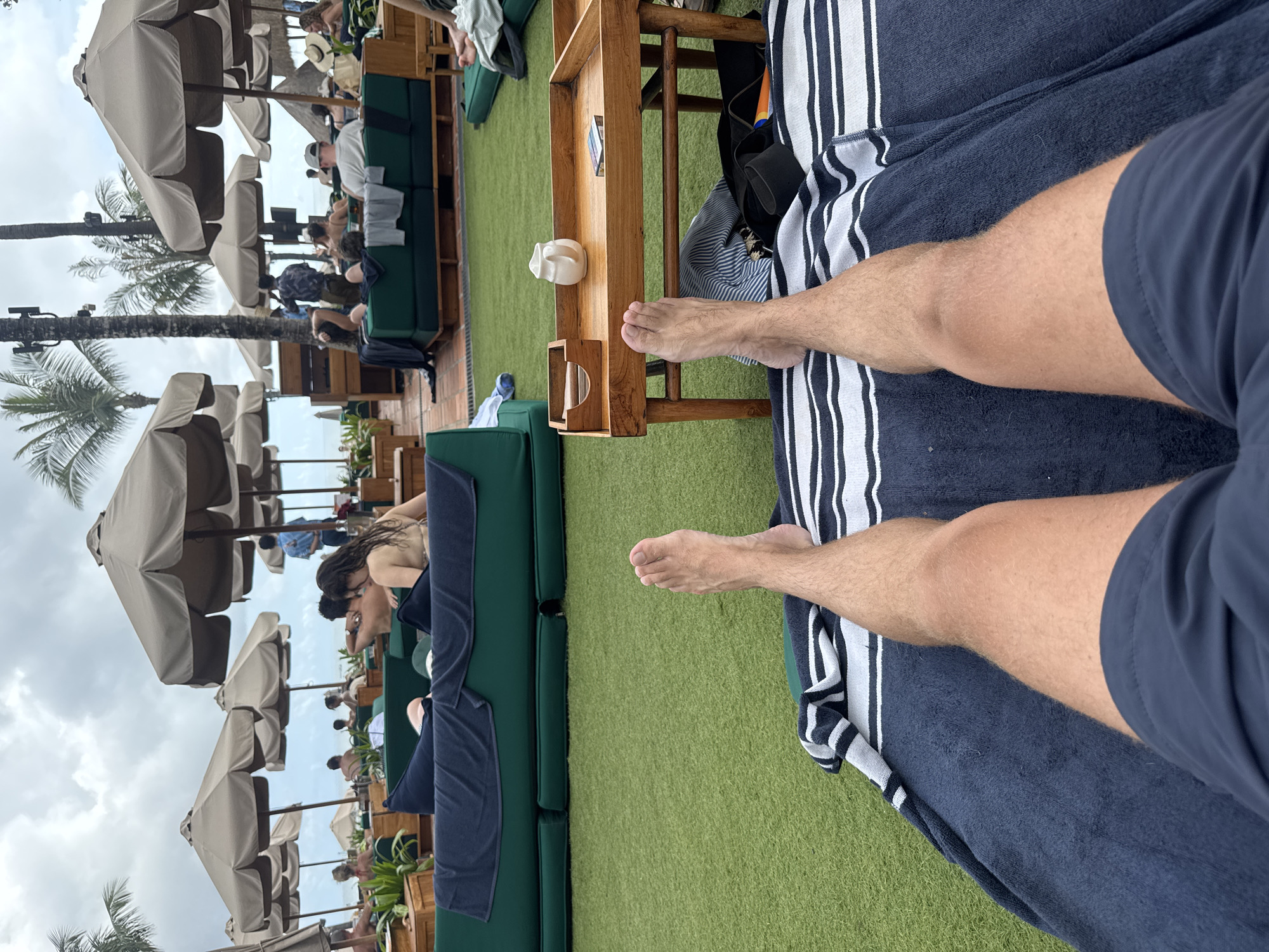 The image captures a scene in North Kuta, Indonesia, with a first-person view of a person reclining on a blue and white striped towel. The person is wearing navy blue shorts and their bare feet are visible, extending towards a small wooden table. The table holds a white ceramic object and a container.
Beyond the table and towel, there is a green artificial turf area where several lounge setups are arranged. These include dark green couches and wooden frames. People are seen relaxing on the couches, and the background features rows of beige umbrellas and palm trees, indicating a resort or beach club setting. The sky is overcast.