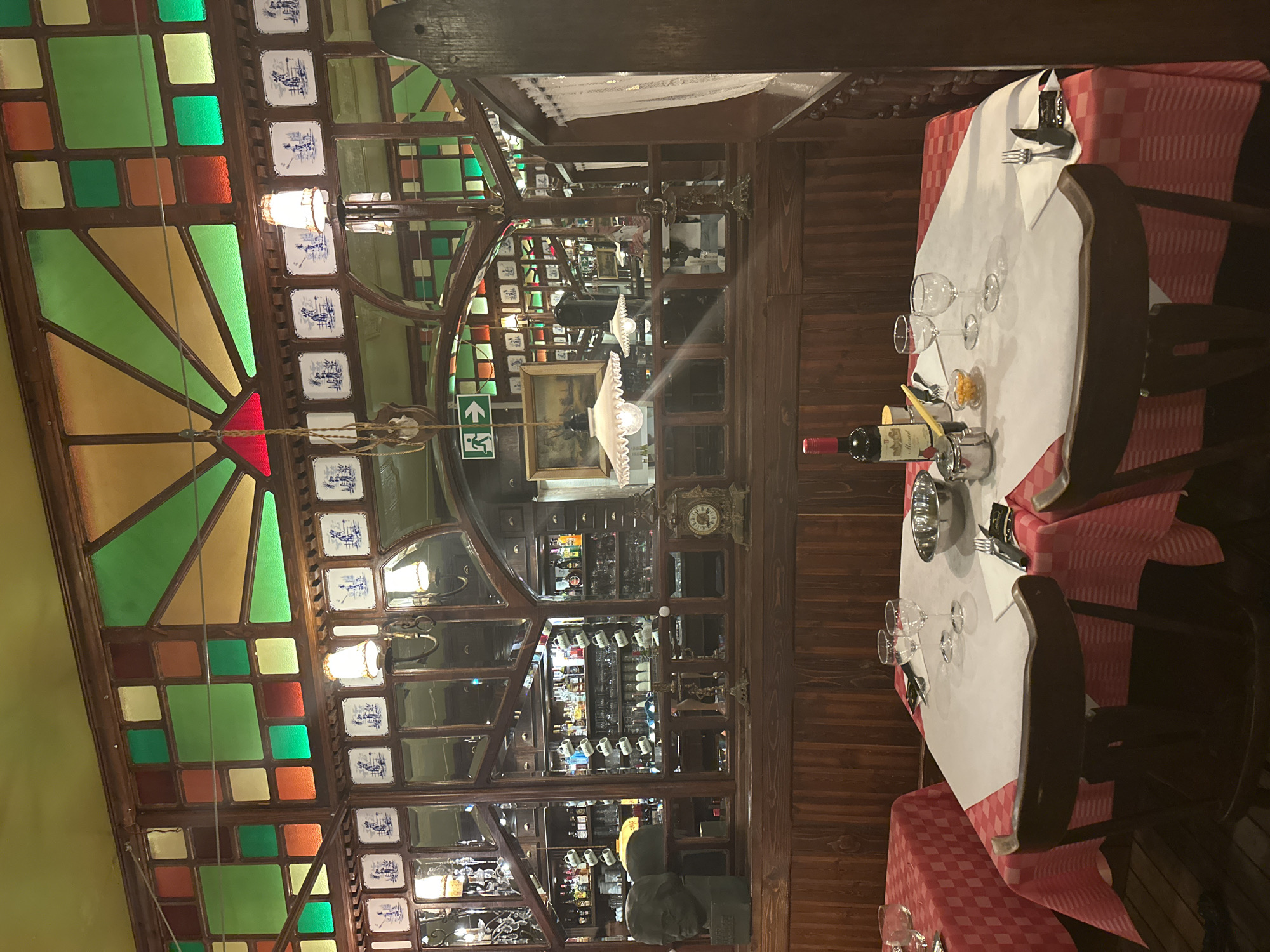 The image depicts the interior of a dimly lit, traditional Belgian restaurant, likely in Bruges, Belgium. The scene is set with wooden paneling and stained-glass windows featuring geometric patterns in green, yellow, and red. In the foreground, two tables are set for dining, each covered with a red and white checkered tablecloth and a white overlay. One table has two wine glasses, silverware, a metal bowl, and a bottle of red wine. The other table, partially visible, also has table settings.
Behind the tables, a wooden bar area is visible with shelves stocked with bottles and glasses. A large mirror reflects the bar and the dining area. Decorative elements include ceramic tiles with blue and white patterns above the mirror, a vintage clock, a bust of a person, and various lamps. A person, possibly a waitress, is visible in the background near the bar. The overall atmosphere suggests a cozy, old-fashioned establishment.