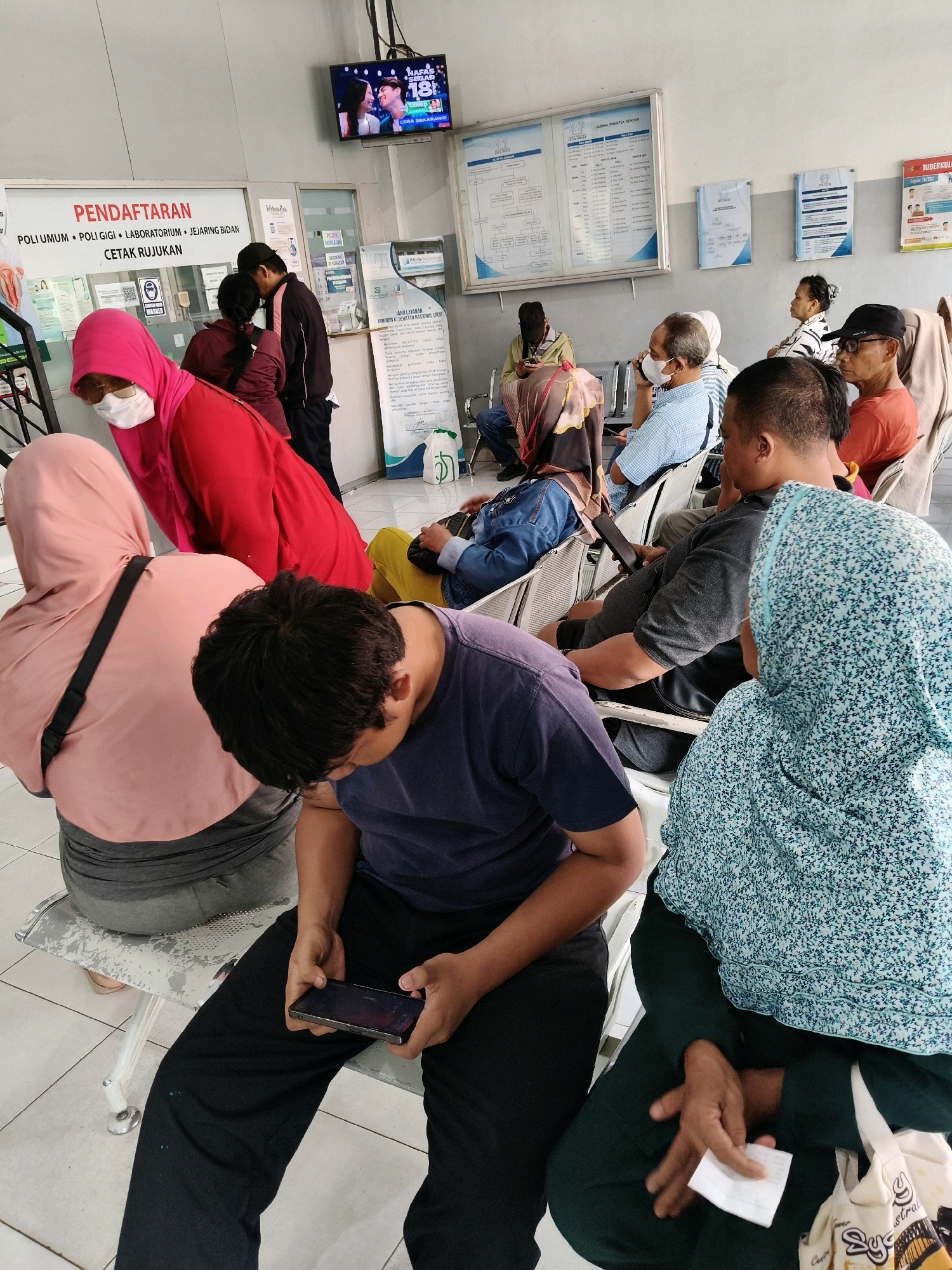 The image depicts a waiting area within a clinic or hospital located in Bekasi, Indonesia, as indicated by text visible on a sign above the entrance that reads "Pendaftaran" (Registration) and other medical-related terms. A group of individuals are seated on a long, metallic bench, waiting for their turn. The environment appears to be indoors, with tiled flooring and administrative signage on the walls.
In the foreground, a young person wearing a navy blue t-shirt is engrossed in their mobile phone. To their right, a woman in a light blue and white patterned hijab sits with her hands clasped. To the left of the young person, another individual wearing a pink hijab and a white mask is partially visible.
Further back, several other people are seated. A man in a blue checkered shirt is on his phone. Another man in a dark grey t-shirt is also present, as is a man in an orange t-shirt wearing glasses and a black cap. In the background, a television screen displays a program. Several informational posters and charts are mounted on the walls, including a schedule of doctor's appointments. The overall scene suggests a typical day in a public health facility.