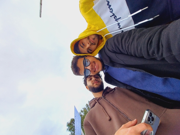 The image displays three young adult males captured from a low-angle perspective against an overcast sky, in what is identified as Al-Muhajerin Municipality, Syria.
The individual on the far left, partially visible, wears a brown zippered hooded sweatshirt. Their right hand, holding a silver smartphone with a multi-lens camera array, is extended into the lower left foreground, indicating they are operating the camera. This subject has a beard and looks directly at the camera with a neutral expression.
The central figure, fully visible from the shoulders up, wears a black jacket over a blue garment. Dark sunglasses obscure their eyes. This individual has dark, curly hair, visible dental braces, and is smiling towards the camera.
The individual on the far right, partially visible, wears a yellow, white, and dark blue color-block hooded sweatshirt with "Champion" lettering and white drawstrings. This subject has facial hair and exhibits a slight smile while looking towards the camera.
The background primarily consists of a uniformly light grey, overcast sky. To the far left, a portion of a blue solar panel array is visible, situated near green foliage. To the far right, a thin, light-colored pole extends vertically into the upper right corner of the frame. The scene suggests an outdoor, informal gathering or selfie.