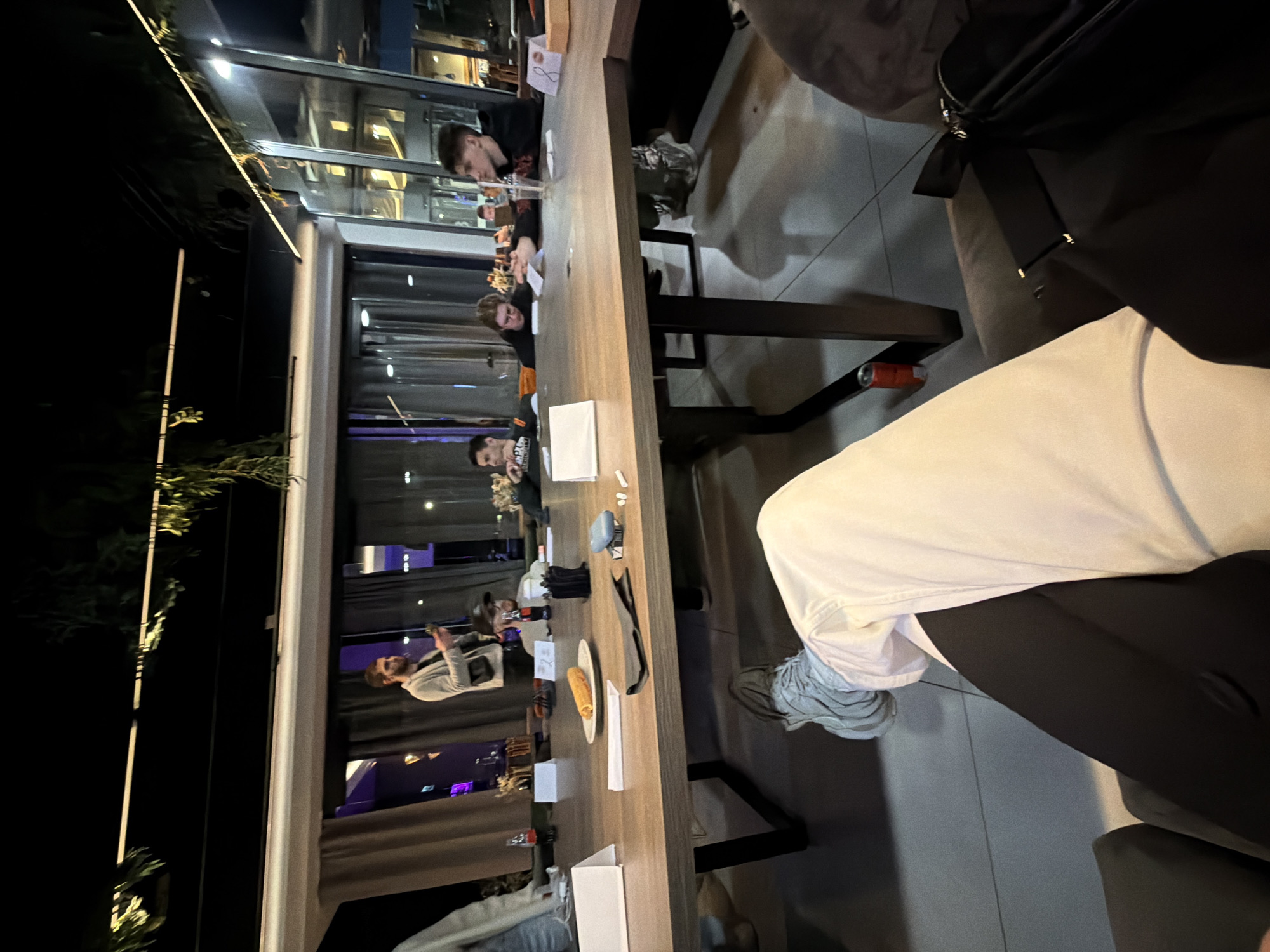 A horizontal indoor shot depicts a group of people seated at a long wooden table. Visible individuals are engaged in various activities, some appearing to be eating or conversing. The table is set with plates, cutlery, and small white boxes.
The setting is a well-lit restaurant or bar, possibly with an outdoor extension, indicated by a glass partition and outdoor vegetation visible to the left. The scene is taken at night, evidenced by the darkness outside and the interior lighting. The location of the city is unknown.