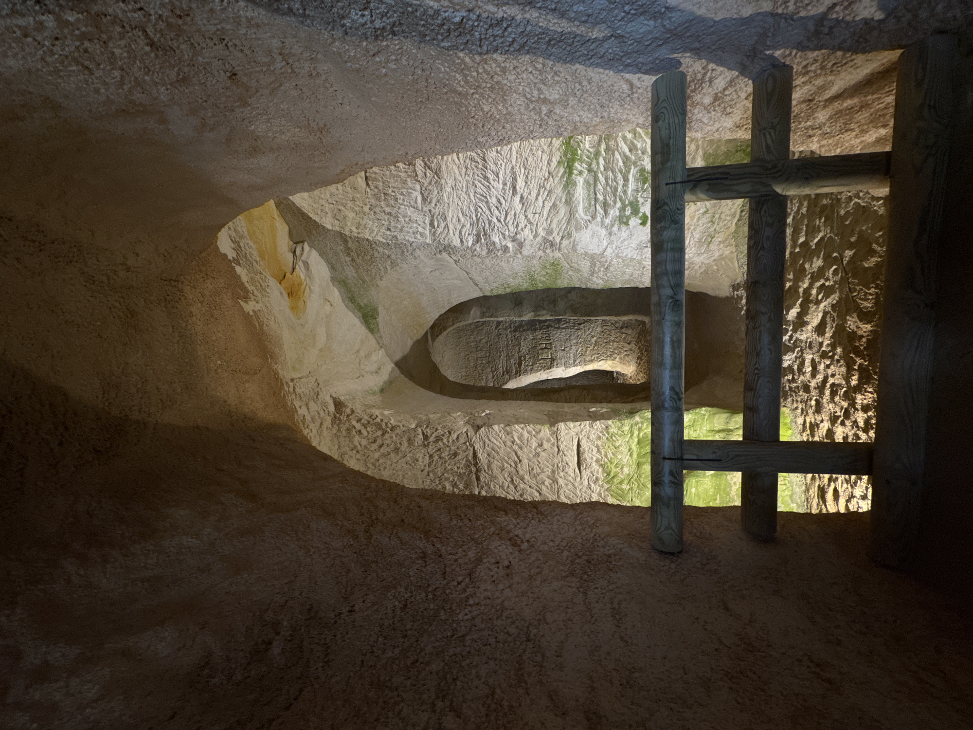 The image shows an interior view of the Piusa Sand Caves in Estonia. The scene depicts a man-made tunnel carved into sandstone, with arched ceilings and rough, textured walls. Natural light, possibly from an opening or artificial lighting, illuminates the space, casting shadows and highlighting the striations in the rock. A wooden railing is visible in the foreground, suggesting a designated pathway or viewing area for visitors. The overall impression is one of subterranean architecture, emphasizing the geological formation and human excavation of the caves.