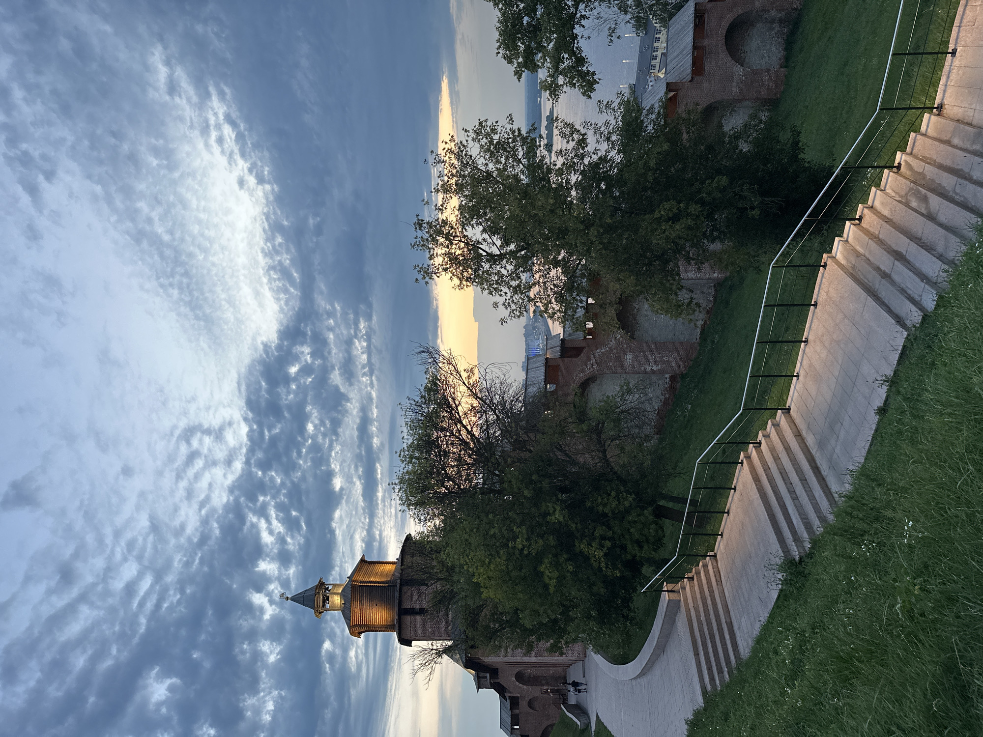 The image depicts a view of the Nizhny Novgorod Kremlin at dusk. In the foreground, a wide, illuminated stone staircase with black railings ascends a grassy hill. To the left of the stairs, a paved pathway curves away, leading towards the Kremlin's brick wall and a wooden tower. The tower is lit from below, highlighting its wooden structure and spire. Several trees with green foliage are positioned between the tower and the pathway. In the background, the wide Volga River is visible, with distant city lights on the opposite bank. The sky is filled with dramatic, textured clouds, transitioning from the pale yellow and orange hues of sunset near the horizon to darker blues and grays overhead. Two figures are walking on the pathway leading to the Kremlin. The scene is characterized by the warm glow of artificial lights contrasting with the cool tones of the twilight sky.