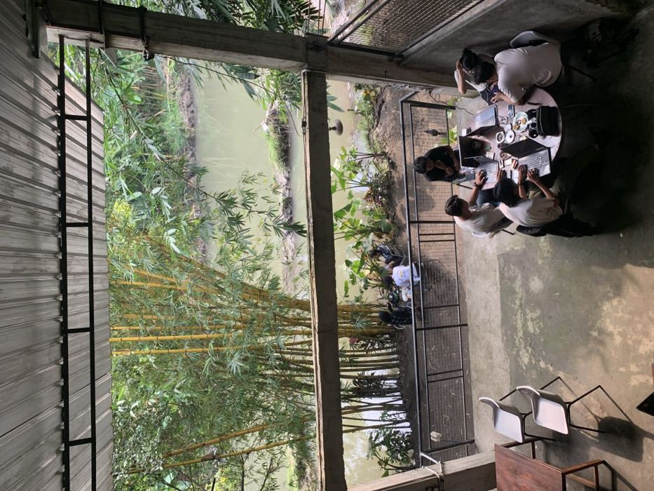 A group of five young adults sit around a round table on a concrete patio, working on laptops. The patio is enclosed by a black metal railing. Beyond the railing, a small river runs through a lush, green landscape. The view of the river is from an upper level, through a covered open area with a corrugated metal roof. There are bamboo plants growing along the river bank. The scene is located in Kecamatan Piyungan, Indonesia, at latitude -7.825, longitude 110.453.