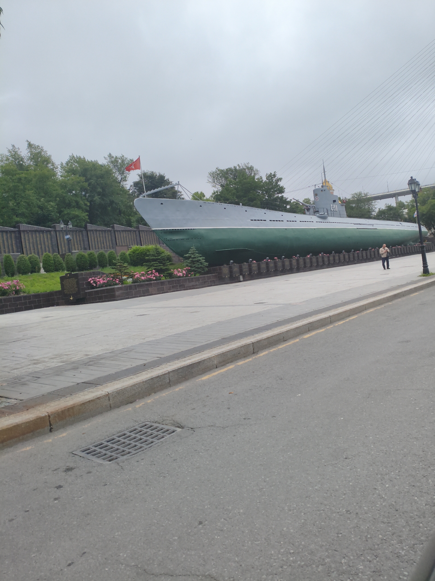 The image depicts a historical submarine, the S-56, displayed as a monument in the city of Vladivostok, Russia. The submarine is positioned on land, with its hull painted in dark green and gray. A red flag is visible on its mast. In the background, a suspension bridge can be seen, along with lush green trees and a cloudy sky. A person is walking along the promenade in front of the submarine. The foreground shows a paved area with a sidewalk, a street, and a storm drain. The overall setting is that of a memorial park or public space dedicated to the submarine.
