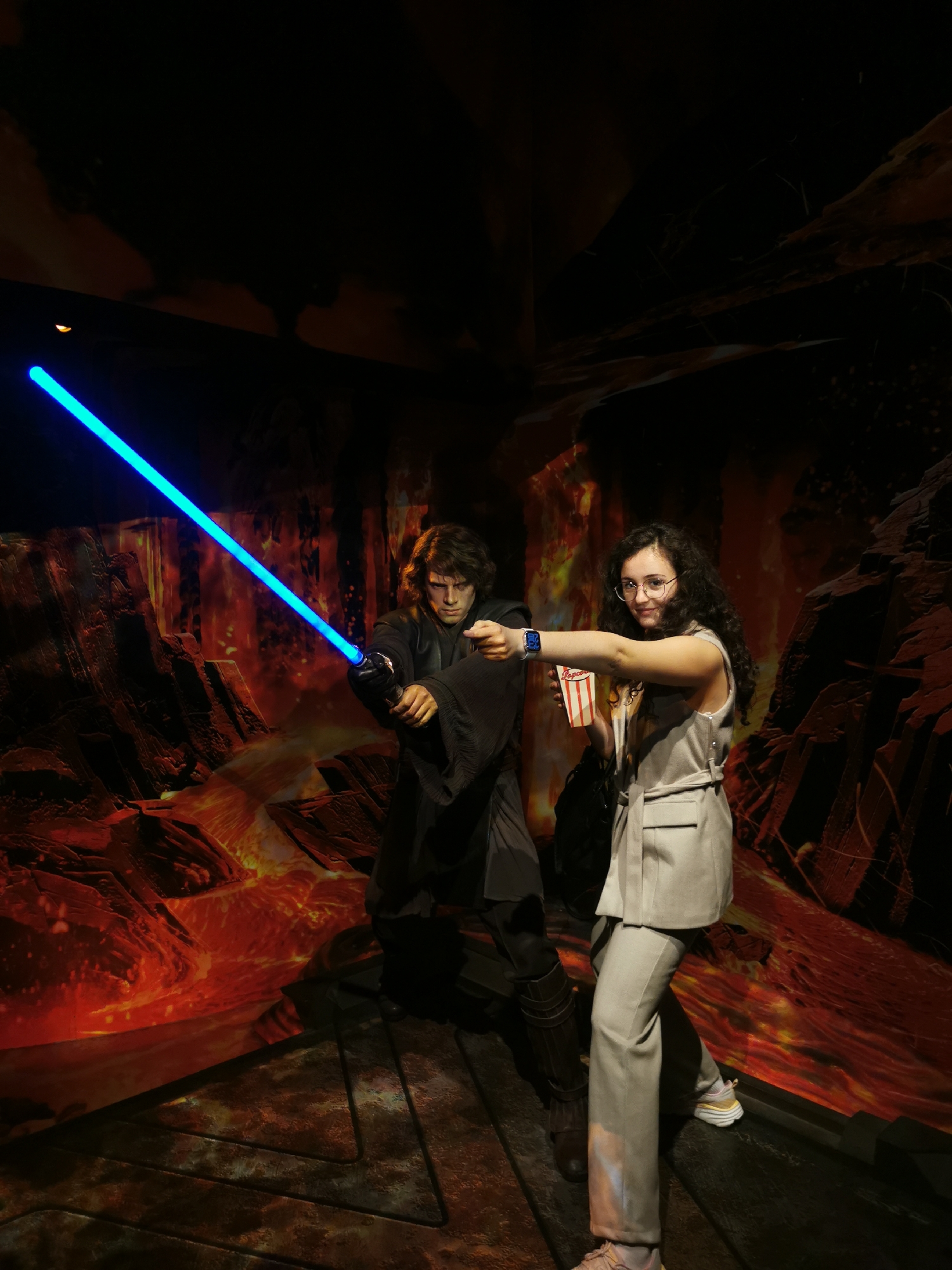 A woman stands next to a wax figure of Anakin Skywalker with a lightsaber, in what appears to be Madame Tussauds in London. The background depicts a lava-filled landscape. The woman is holding a popcorn container.
