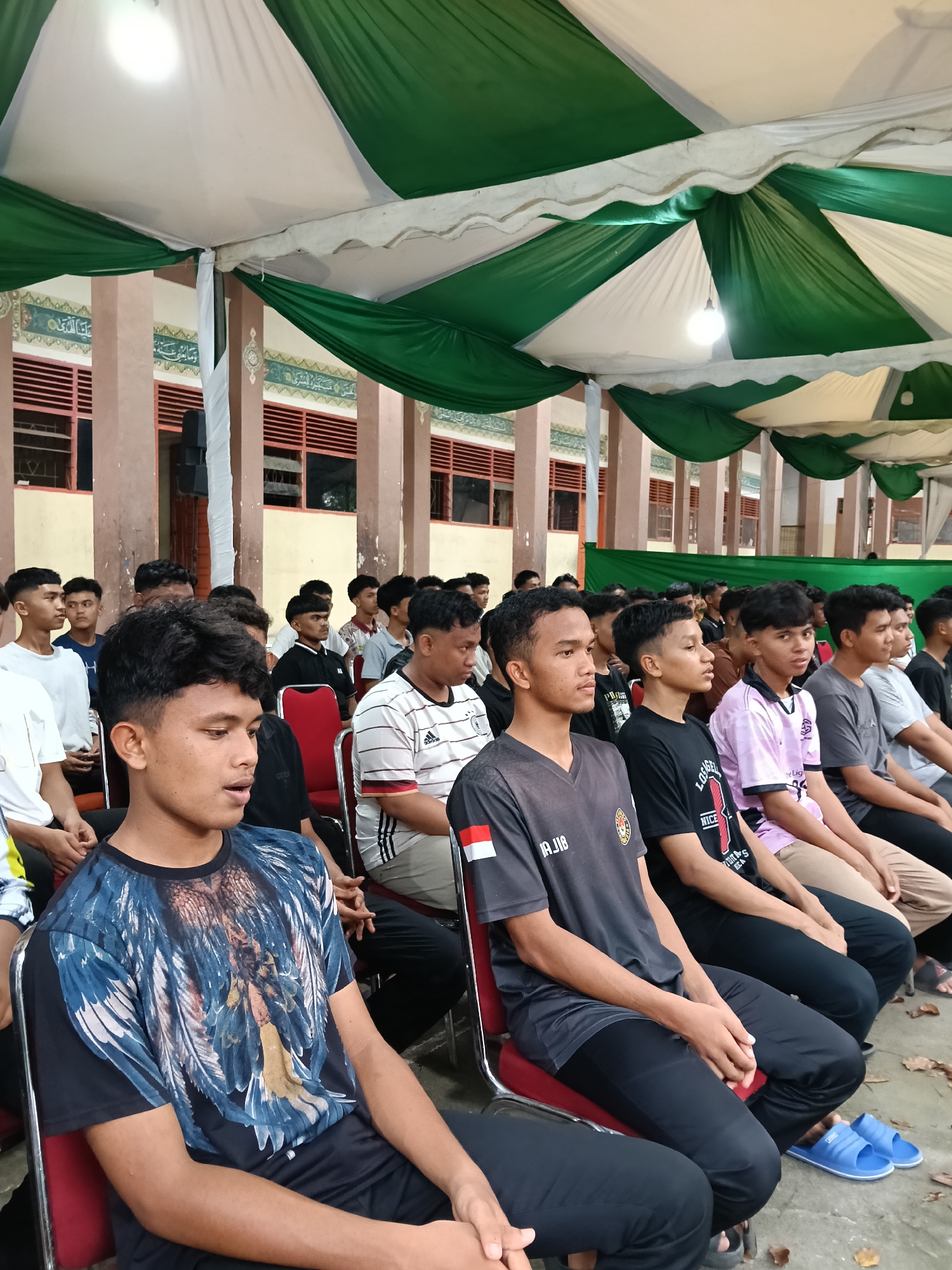 The image shows a gathering of young men seated in rows under a tent-like structure, likely in Lhokseumawe, Indonesia, suggested by the architectural style of the buildings in the background and the general appearance of the subjects. The tent is made of green and white fabric, suspended above. Fluorescent lighting hangs beneath the tent. The men are seated in red plastic chairs, generally looking towards the same direction. The clothing varies, some wearing casual t-shirts and pants, others in athletic apparel. There are mosque or civic buildings in the background with windows, pillars, and architectural motifs common in Indonesian mosques. The overall scene gives the impression of an organized event or meeting.