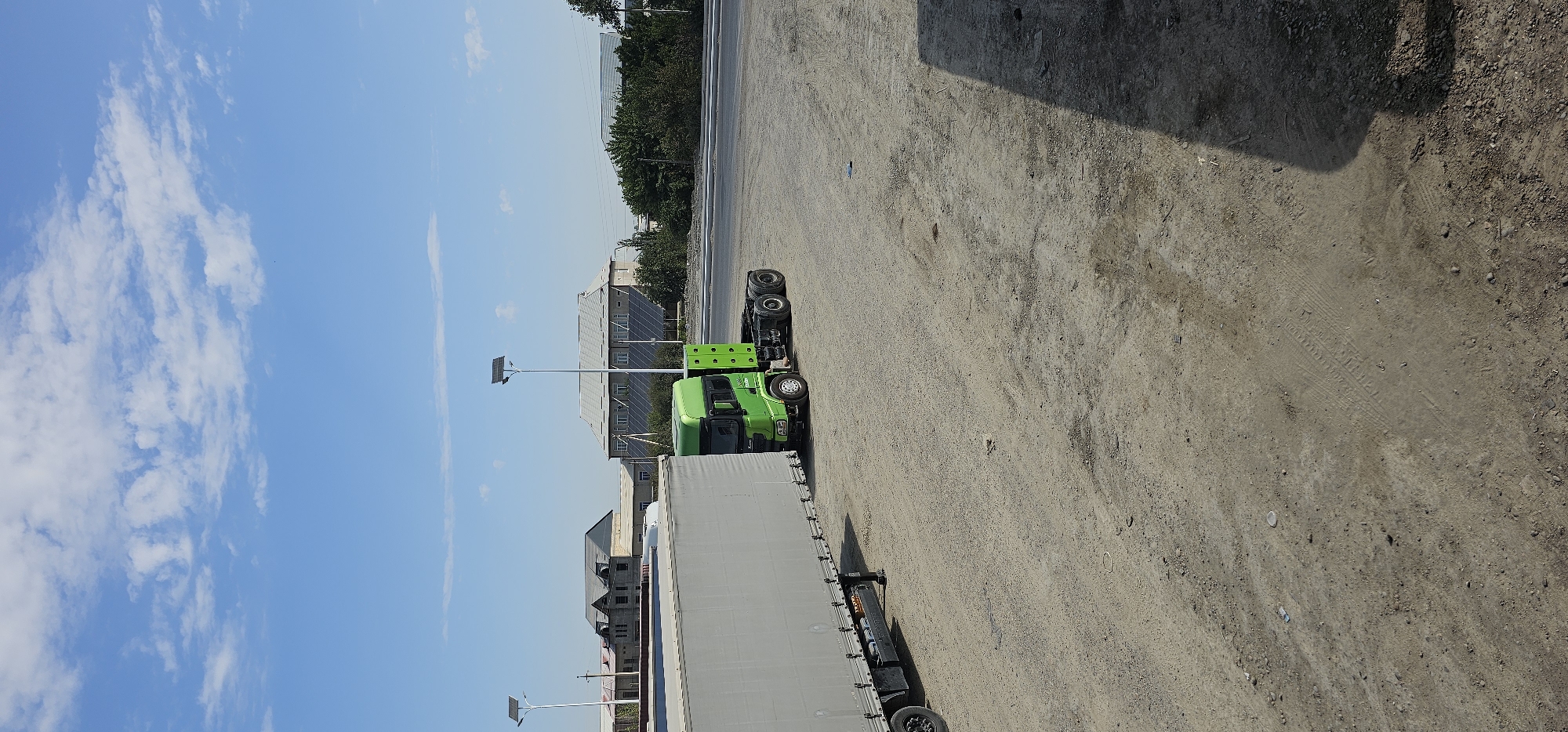 The image captures a daytime outdoor scene featuring a parked green semi-truck with a detached trailer in the city of Sheyhancha, Uzbekistan. The truck, a prime mover without a load, is positioned on a dirt and gravel lot. To the left of the truck, a large, light-colored trailer is parked. In the background, several buildings are visible, including structures with light-colored roofs and some under construction. Two modern lampposts with solar panels stand in the mid-ground. The sky is clear blue with scattered white clouds. The ground surface is predominantly unpaved, with visible tire tracks.