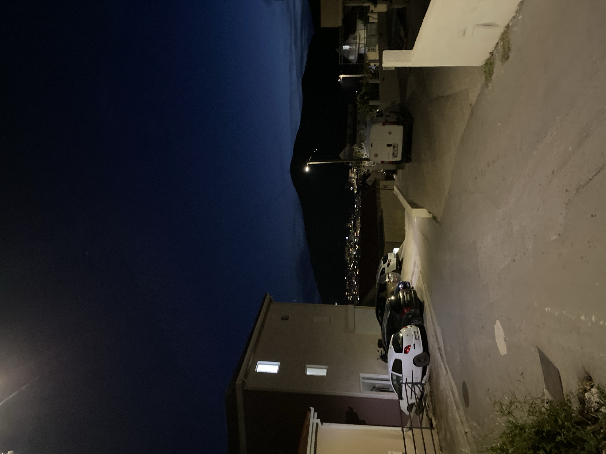 The image shows a night scene in Budva, Montenegro. A narrow street lined with buildings and parked cars slopes downward. The street is lit by streetlights, and the lights of the city can be seen in the distance against a dark sky and mountainous backdrop. The architecture is a mix of residential buildings. A row of parked cars is visible on the right side of the street, including a white car parked at the front. Vegetation is visible at the lower right corner of the image. The sky is dark blue with a hint of purple near the horizon.