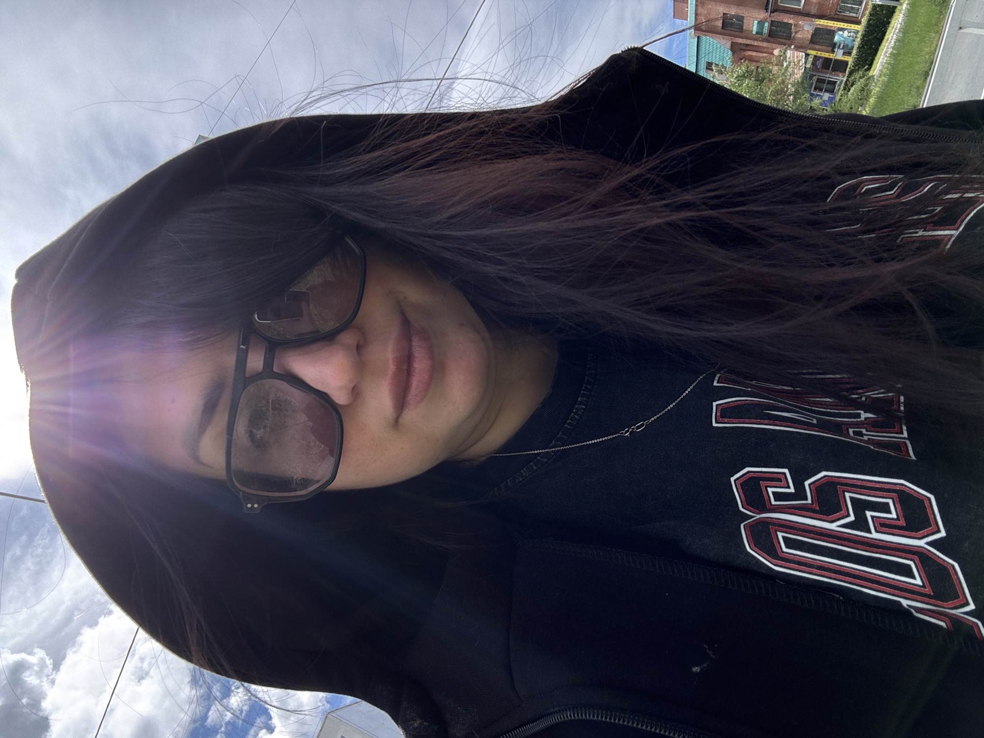 A person with long, dark hair and black-framed glasses is the central subject of this close-up, angled outdoor shot. They wear a faint, pleasant smile with lips slightly parted. The individual is dressed in a dark hooded jacket over a dark t-shirt, which is adorned with a thin silver chain necklace featuring a small, interlocking pendant.
The photograph captures the person from a low, upward-looking angle, possibly a selfie. A strong sun flare emanates from the left side of the frame, indicating bright daytime conditions. The background shows a partially cloudy sky and elements of an urban or suburban setting, including a reddish-brown brick building with multiple windows, green foliage, and a hint of a street or sidewalk.
On the front of the person's dark t-shirt, stylized letters in red and white outline are visible. The letters "SS" are clearly discernible, suggesting a logo or part of a word. The overall mood of the image is casual and relaxed, emphasized by the bright sunlight and gentle expression.