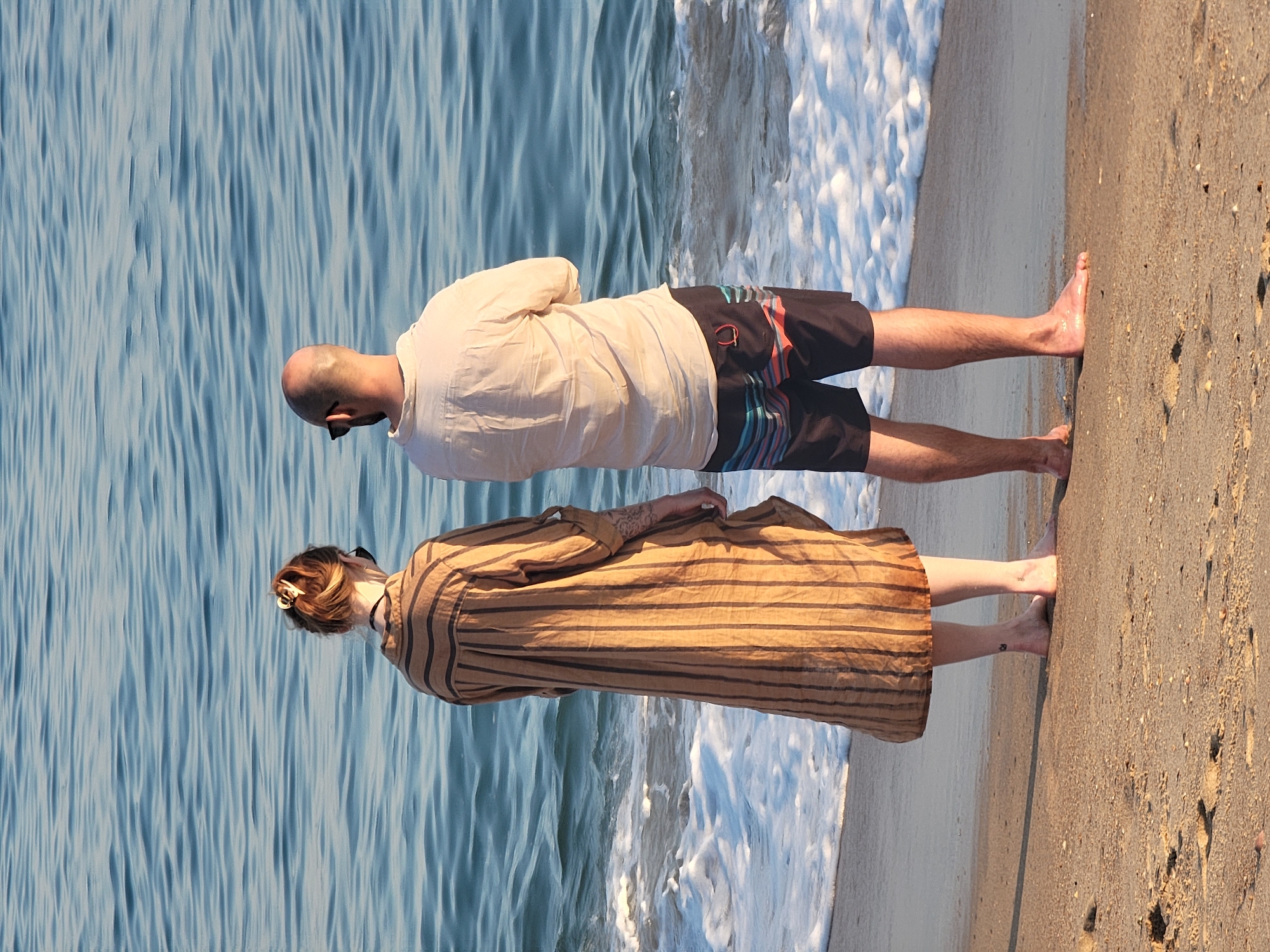 Two individuals, a woman and a man, stand facing the ocean on a sandy beach, likely in Quarteira, Portugal, given the context. The woman, on the left, wears a long, striped brown and black dress. Her hair is pulled back, and she is wearing sunglasses. The man, on the right, is bald and wears a light-colored, short-sleeved collared shirt and black swim trunks with multi-colored stripes. Both are barefoot, with their feet on the sand. Gentle waves are breaking on the shore in front of them. The ocean is a vibrant blue with horizontal ripples. The overall scene is calm and serene, suggesting a moment of quiet observation of the sea.