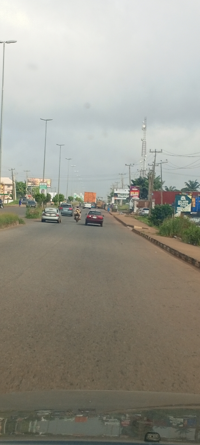 The image displays a daytime street scene on a multi-lane asphalt road in Akure, Nigeria, under an overcast sky. The perspective is from inside a vehicle, indicated by a dashboard reflection across the bottom edge.
Traffic is moving forward in the visible lanes. In the left lane, a silver sedan with an open trunk, containing white items, is followed by a dark blue or grey sedan. A yellow and black motorcycle with two occupants is positioned between the lanes. In the right lane, a red sedan is followed by a truck carrying a large orange shipping container. Behind this, a yellow construction vehicle is visible.
The left side of the road features a series of tall streetlights, power poles, and partially obscured buildings behind vegetation, with multiple billboards visible above.
The right side of the road includes additional power poles, a prominent telecommunication tower, and dense vegetation. Commercial establishments are present, characterized by multiple shipping containers, some dark red/brown and others blue. One blue container prominently displays "AYA SUPER HOTEL AKURE." Further billboards are visible. A single pedestrian in a blue outfit is observed walking on the far left sidewalk, away from the camera. Other indistinct figures are present on the right sidewalk. The asphalt road surface exhibits wear.