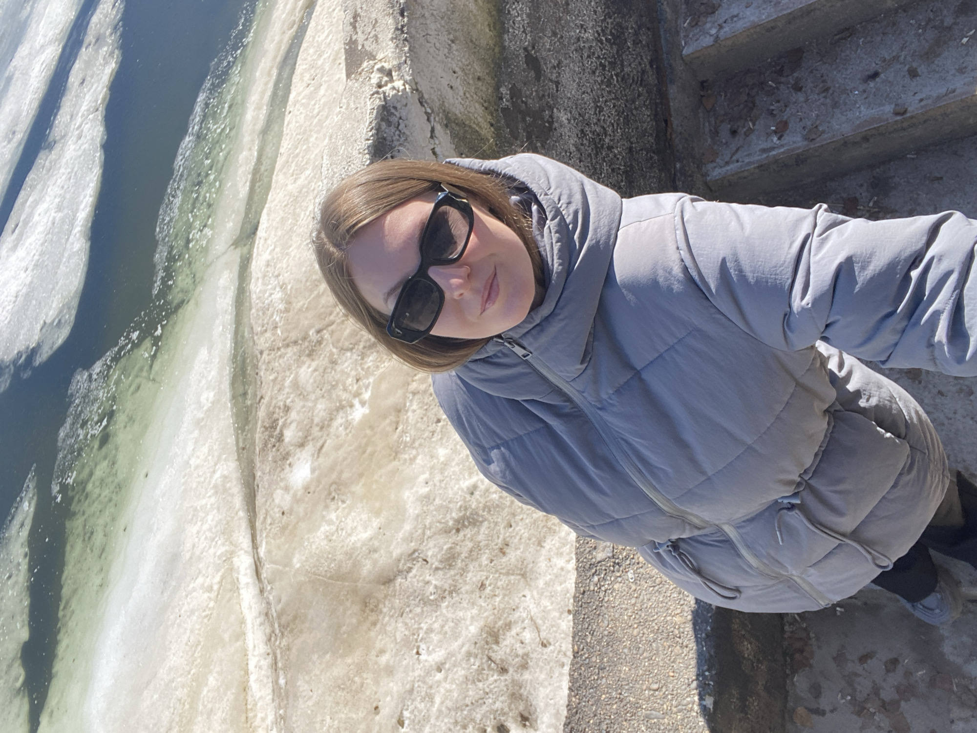 A young woman wearing a gray puffer jacket and sunglasses is taking a selfie outdoors. She is standing on a concrete structure, possibly steps or a retaining wall. To the left of the frame, there is a body of water with ice floes, suggesting a cold climate. The location is Chaikovsky, Russia.