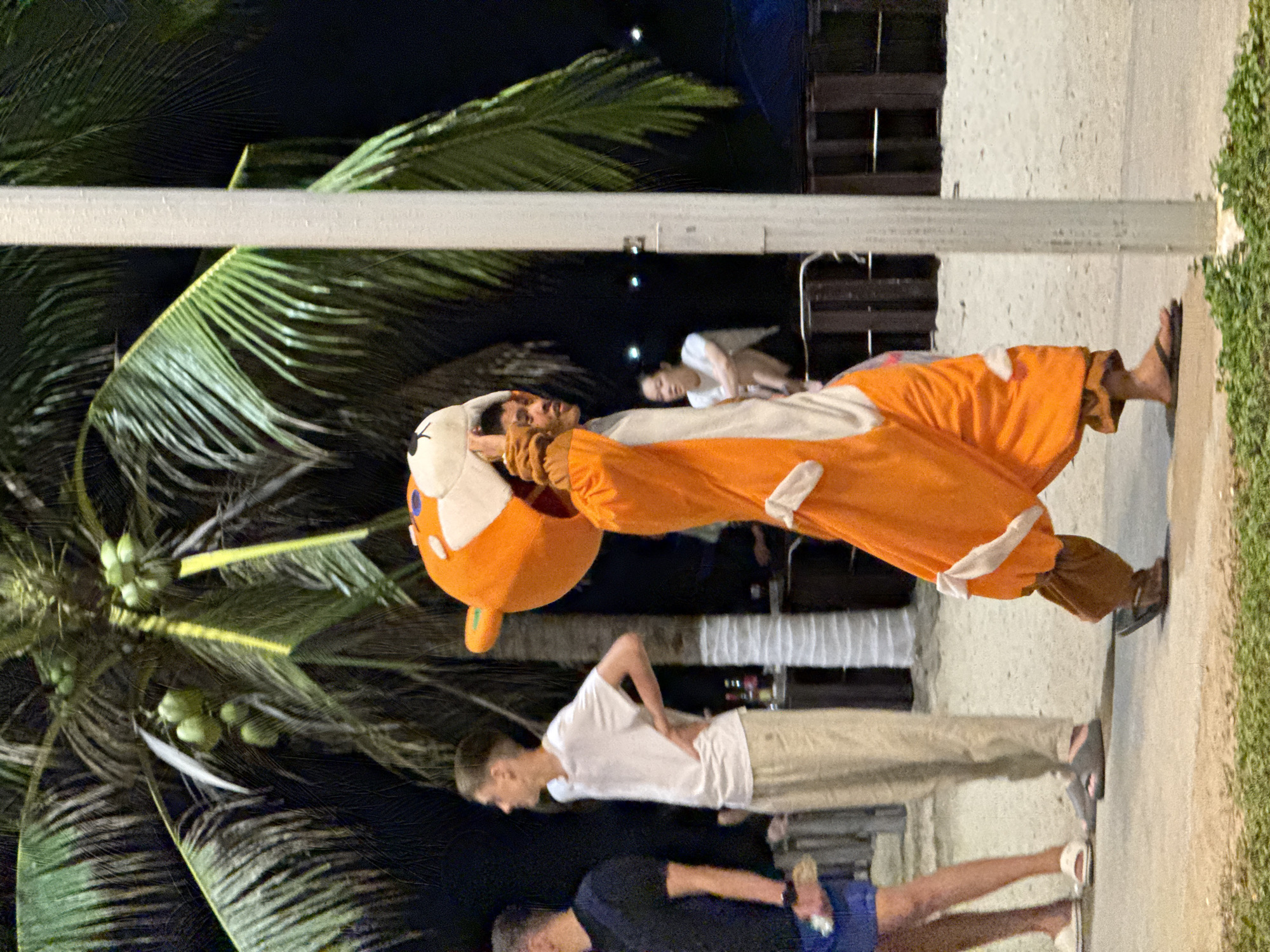 Captured in the Nha Trang Ward, Vietnam, the image features a person wearing a full-body costume of an orange and white cartoon animal, potentially a fox or dog, standing on a concrete pathway. The individual is surrounded by others, some dressed in casual clothing, including a man in a white shirt and tan pants, and another in a black shirt with blue shorts. A tall coconut tree is visible on the left side of the frame, its leaves framing the scene. The background hints at a nighttime setting, with faint lighting and indistinct structures, possibly buildings or temporary setups. The overall impression is that of a street performance or public event.