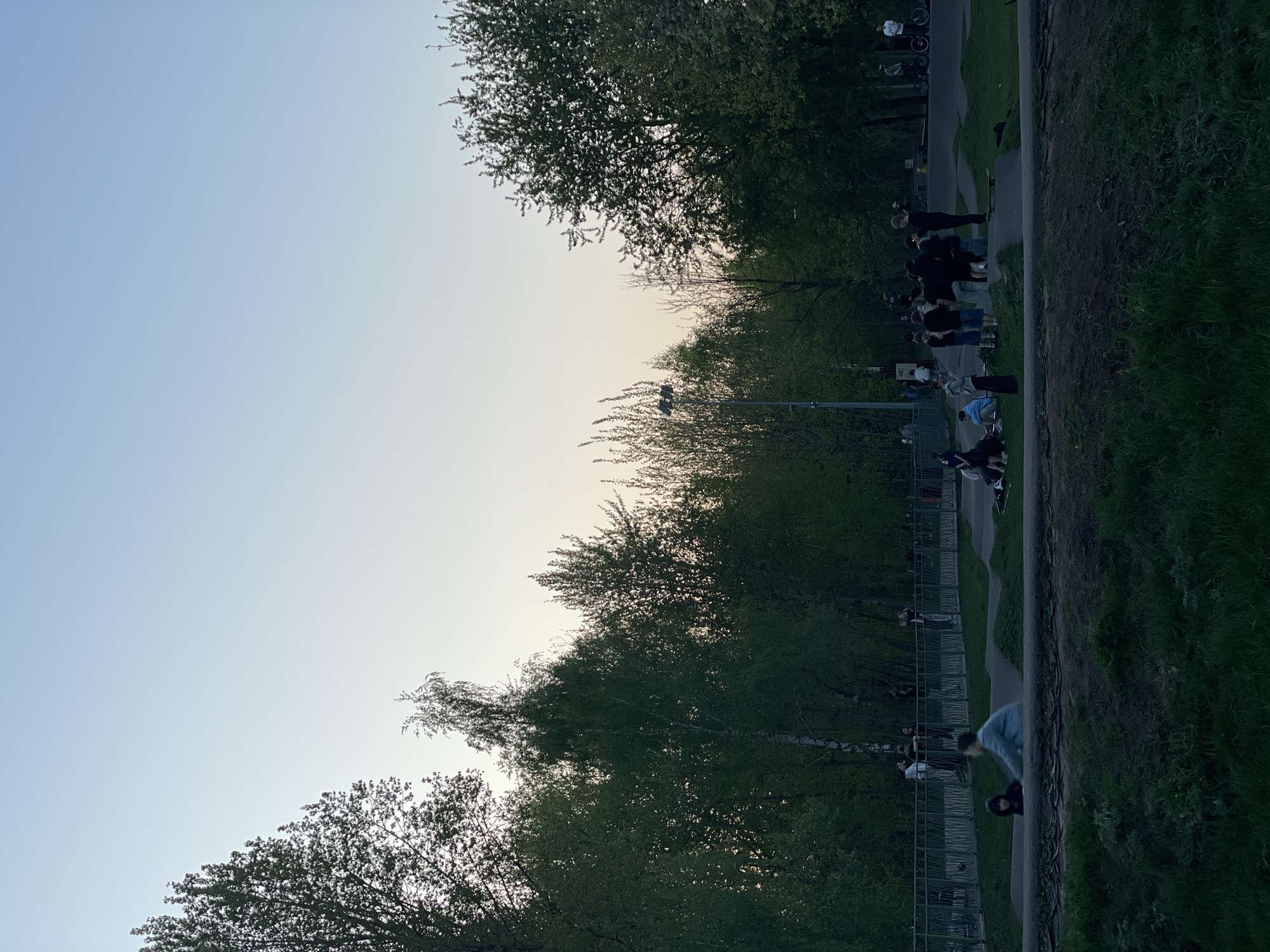 The image shows a park scene at dusk. A group of people is congregated along a pathway bordered by trees and grass. A metal fence separates the pathway from the trees. A streetlight is visible amidst the trees. The sky is light blue and appears to be close to sunset. The location is Kazan, Russia.