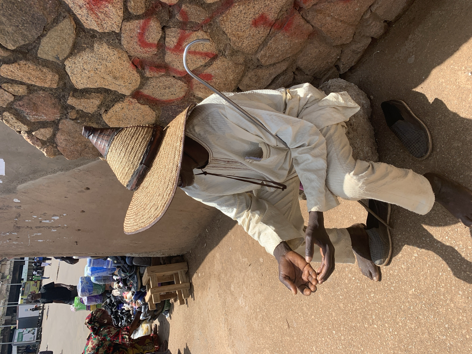 In Jos, Nigeria, an elderly man sits in the shade of a large, woven conical hat with a dark leather brim. He is dressed in loose-fitting, off-white traditional attire. His hands are open and resting on his lap, and a curved metal cane lies beside him. He is seated on a large rock against a wall constructed of rough-hewn stones, some of which are marked with red paint. The ground is a dusty, reddish-brown earth, and in the background, a busy market scene unfolds. Stalls display various goods, including stacks of colorful plastic containers and piles of footwear. People are seen browsing and interacting within the market. The overall scene depicts a moment of rest amidst the activity of a Nigerian marketplace.