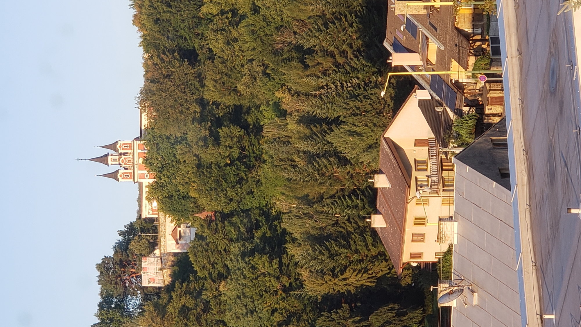 The image is an outdoor, eye-level shot of a cityscape with a prominent building featuring multiple spired towers on a hillside covered in trees. The building, likely a church or monastery due to its architecture, is red and white with dark spires. In the foreground, there are several residential buildings with red-tiled roofs, some equipped with solar panels. A satellite dish is visible on one of the roofs. The environment appears to be a sunny day with clear skies. The scene is likely located in Prešov, Slovakia, given the architectural style and the presence of residential areas bordering a forested hill. There are no people visible in the image.