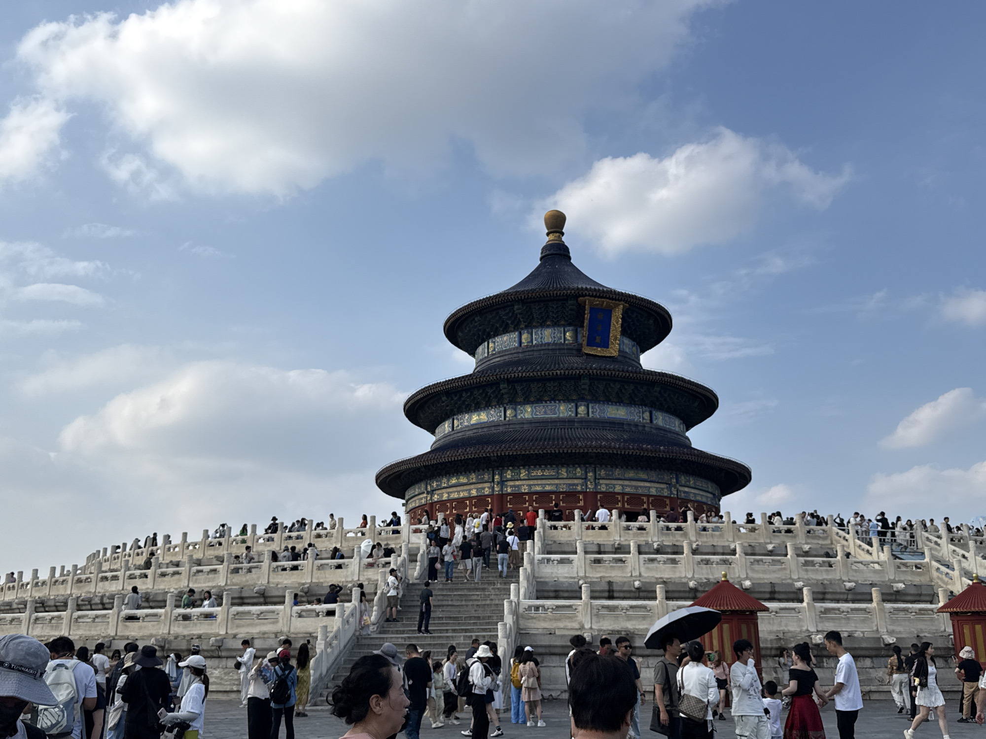 The image captures the exterior of the Temple of Heaven in Dongcheng District, China, under a bright, cloudy sky. The iconic, multi-tiered, circular building with a dark blue roof and intricate gold and blue decorations stands prominently in the background. The building is surrounded by a tiered, white stone terrace filled with numerous people. Visitors, dressed in casual summer attire, are seen walking, standing, and ascending the stairs of the terrace. Some individuals are holding umbrellas, and others are engaged in conversation or taking photographs. The overall scene depicts a busy tourist day at a significant historical landmark.