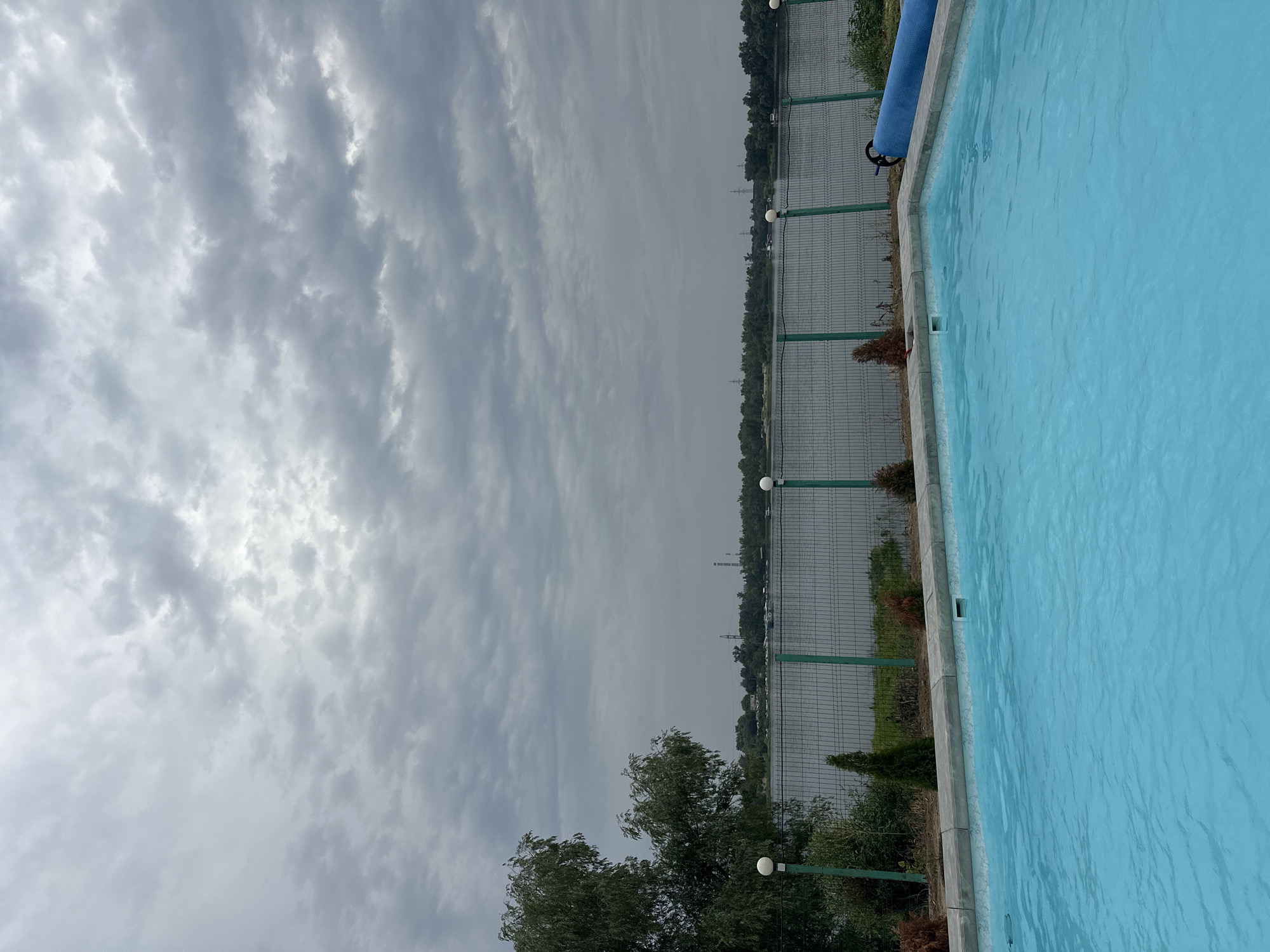 An outdoor swimming pool with bright turquoise water is in the foreground. A grey, overcast sky with textured clouds fills the top half of the image. Beyond the pool, a dark wire fence with green posts runs horizontally across the midground. Behind the fence, a calm body of water, likely a river, is visible. On the far bank of the river, a dense line of green trees stretches across the horizon, punctuated by industrial structures and power lines. A large tree with dark green foliage is on the left side of the image, near the fence. A rolled-up blue cover is positioned on the right side of the pool's edge. The scene appears to be located in Balakovo, Russia, possibly in a recreational area or resort, given the pool and riverfront setting. There are no people visible in the image.