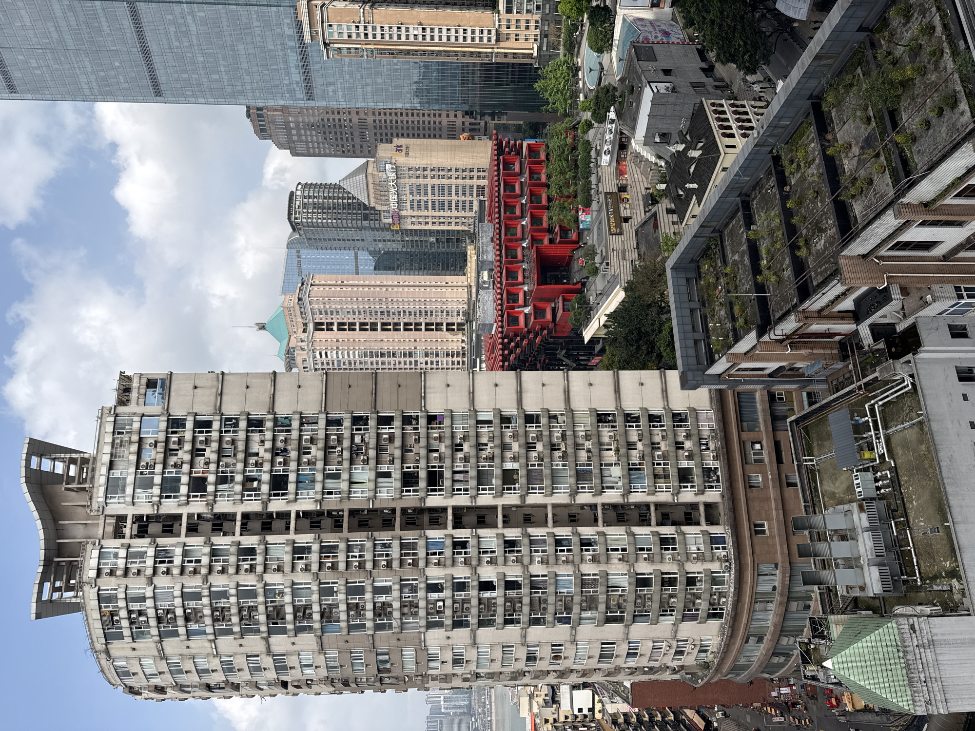This high-angle shot depicts a dense urban scene in Yuzhong District, China. The foreground is dominated by a large, curved, grey concrete building with numerous windows and protruding air conditioning units. Behind this building are a mix of taller buildings of varying heights and architectural styles, including glass skyscrapers, brown buildings, and a distinct red, geometric structure. To the right of the concrete building are rooftops of other buildings, some with vegetation. The sky is partly cloudy with blue patches.