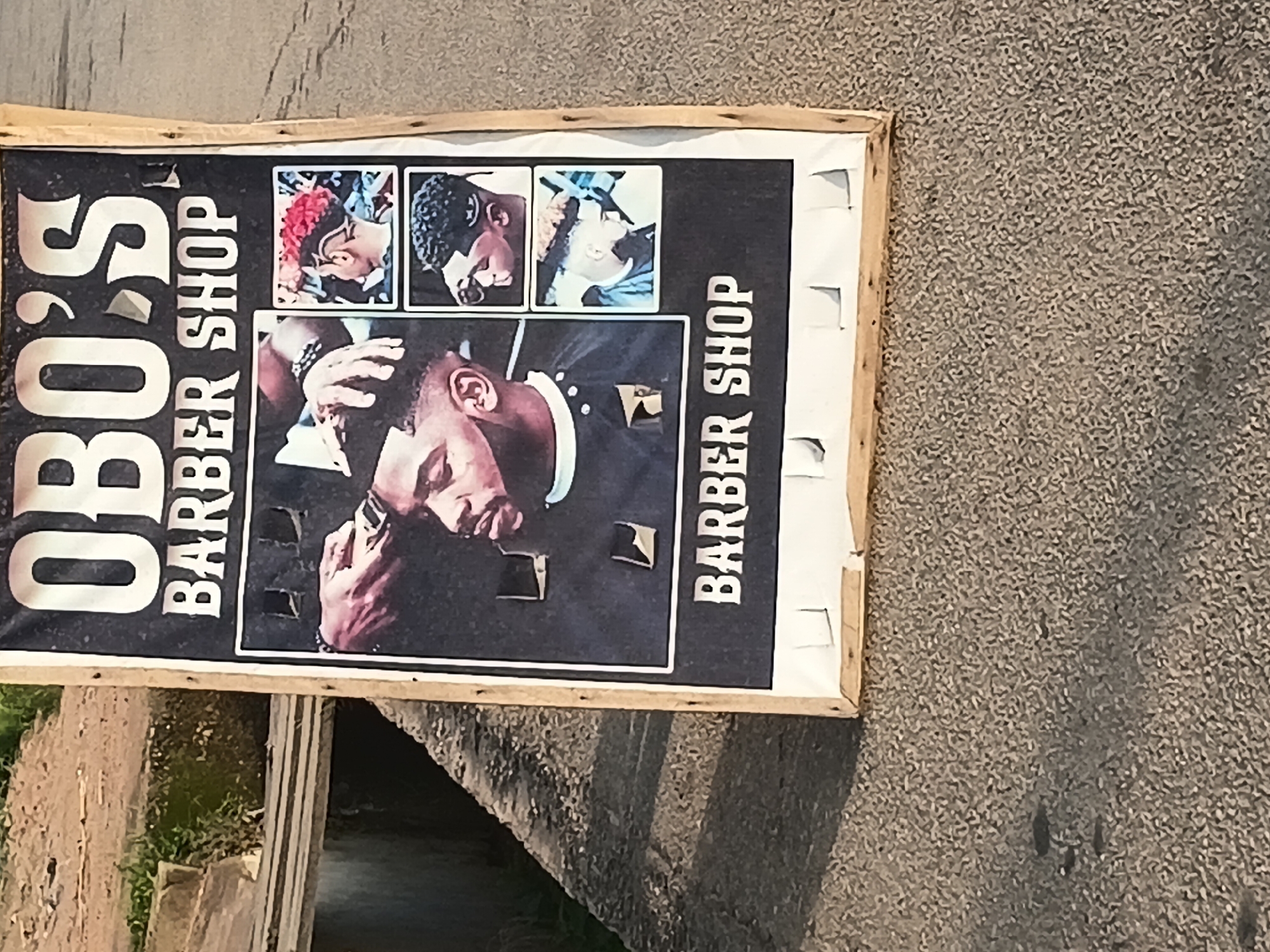 An A-frame sign for "OBO'S BARBER SHOP" stands outdoors on a concrete surface. The sign features a dark background with large white text at the top that reads "OBO'S" and "BARBER SHOP" below. The center of the sign displays three images of men with various hairstyles, with the largest image showing a barber's hands giving a haircut to a man. The sign is framed by light-colored wood and is attached with white tape. The environment appears to be an urban street in Jalingo, Nigeria, with a slightly worn concrete surface and a background that suggests a roadside with some vegetation and a drainage ditch. The lighting indicates it is daytime.