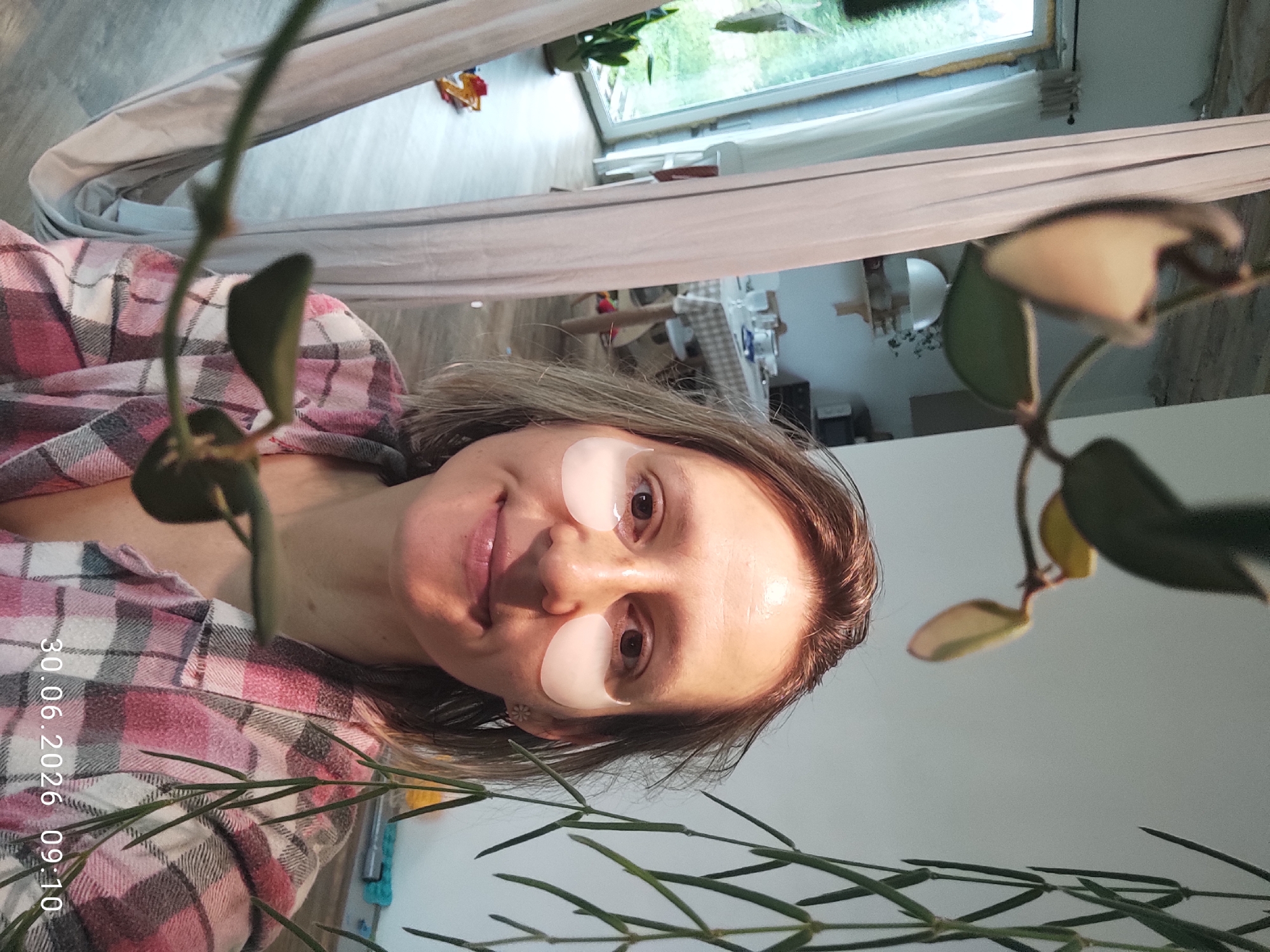 In a domestic setting, a woman with light brown hair and fair skin is positioned centrally in the frame, looking towards the camera. She is wearing under-eye patches and a pink and white plaid shirt. The background reveals a living space with a dining table set in the mid-ground and large windows offering a view of greenery. The foreground is occupied by the blurred branches and leaves of a hanging plant. A timestamp of "30.06.2026 09:10" is visible in the lower right corner. The scene suggests an indoor environment possibly in Malyi Bukor, Russia.